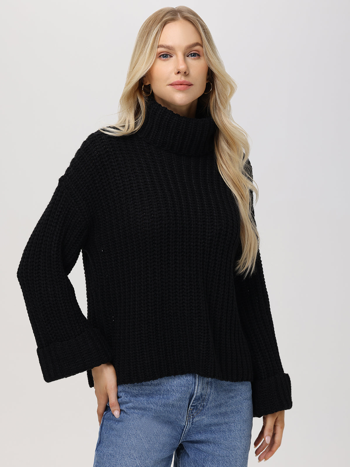 A person with long blonde hair wears 525 Americas cozy Turtleneck Shaker Pullover—a black waffle-textured sweater with oversized sleeves—and blue jeans, standing against a light gray background. They casually slip their left hand into a pocket.