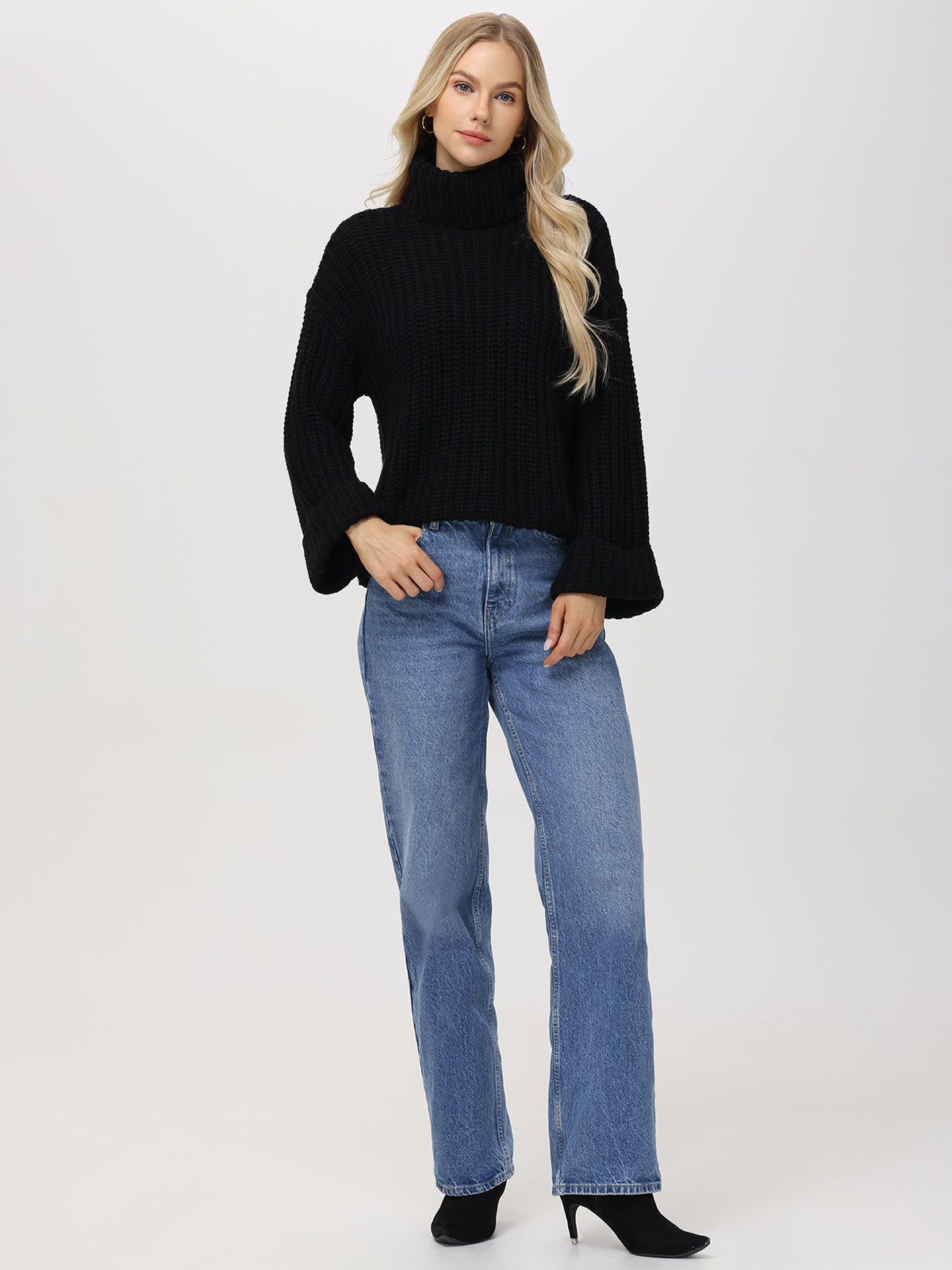 Wearing a 525 America black Turtleneck Shaker Pullover with oversized sleeves, a person with long hair stands against a plain background, paired with blue jeans and black heeled boots, casually resting their hands in their pockets.