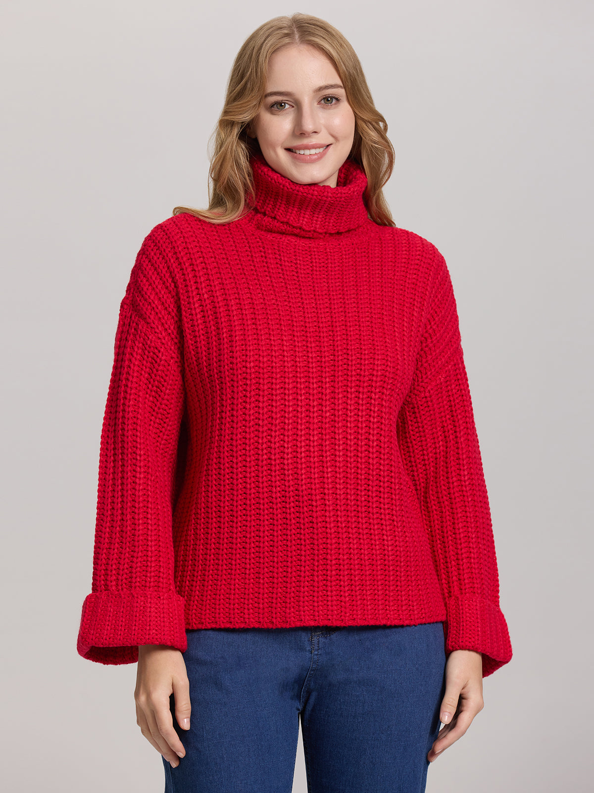 Someone in a red 525 America Turtleneck Shaker Pullover with oversized sleeves stands against a plain background. The textured knit sweater is paired with blue jeans. Their hands rest by their sides, and they have a neutral expression.