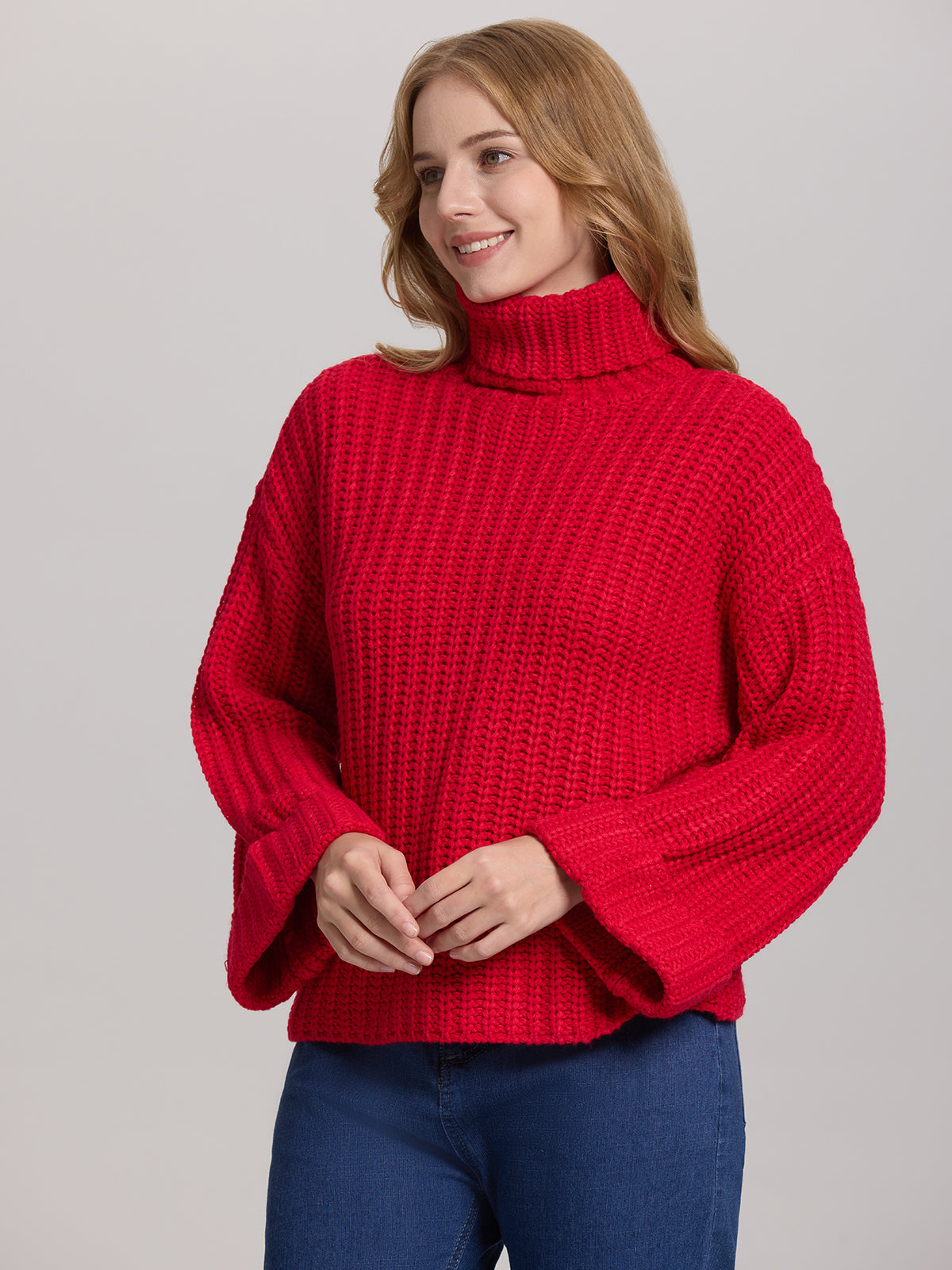 A person with shoulder-length hair wears a cozy turtleneck from 525 America, showcasing an oversized chunky red pullover with roomy sleeves, paired with blue jeans. Theyre posed against a plain gray backdrop, gazing slightly away and sporting a subtle smile.