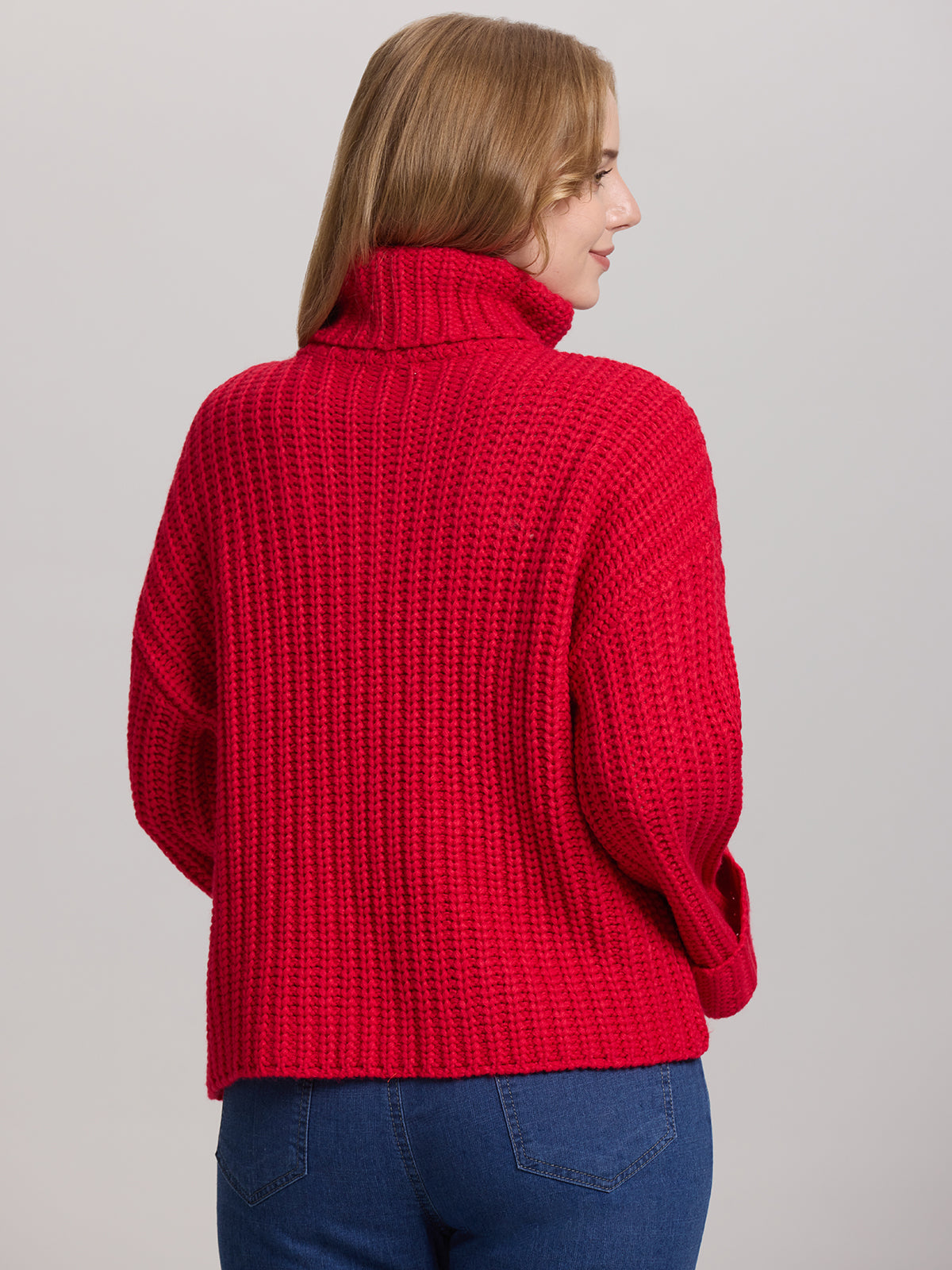 A person with long hair is seen from the back, wearing a cozy 525 America turtleneck with oversized sleeves in a chunky red sweater and blue jeans against a plain background.