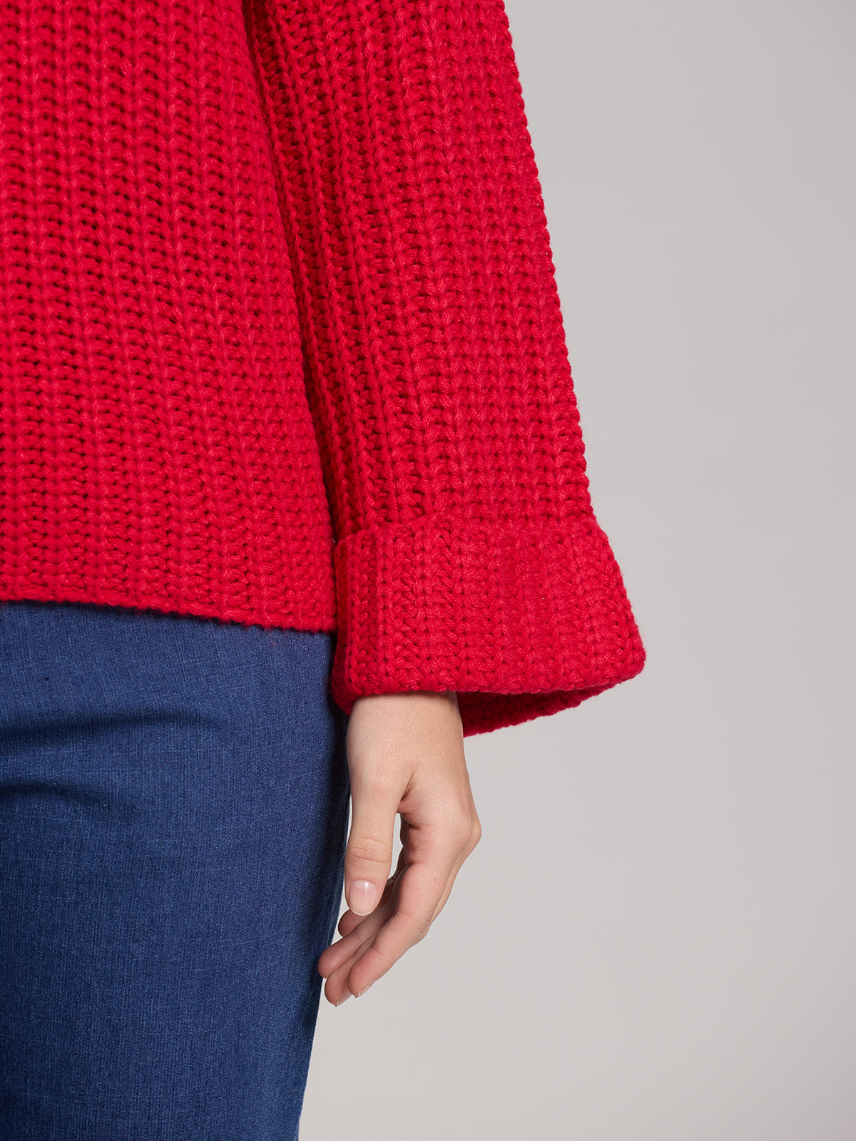 A person wearing a bright red, textured Turtleneck Shaker Pullover by 525 America stands against a plain background. The pullover features oversized sleeves and wide cuffs, showing one side of the sweater and part of their blue pants, with their hand resting relaxed by their side.