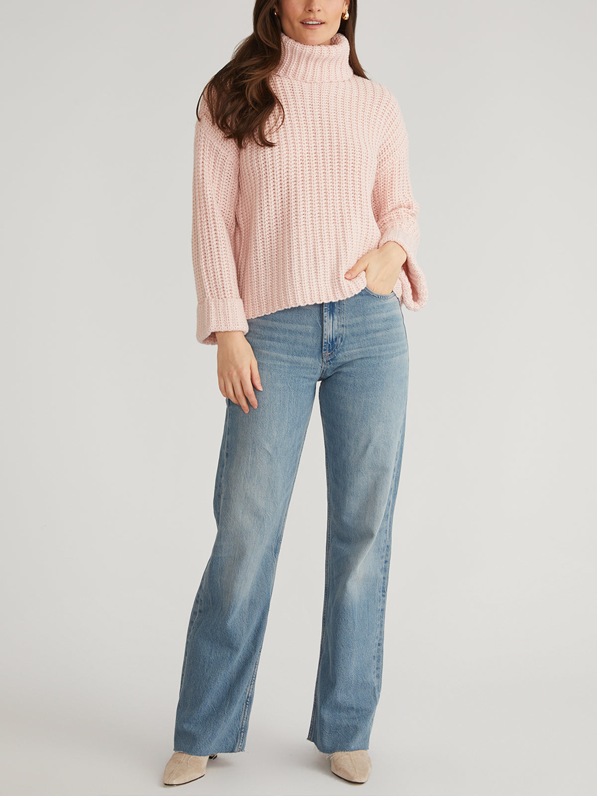 A person stands against a plain background wearing the 525 America Turtleneck Shaker Pullover with oversized sleeves, light blue high-waisted wide-leg jeans, and beige shoes—one hand in their pocket and the other by their side.
