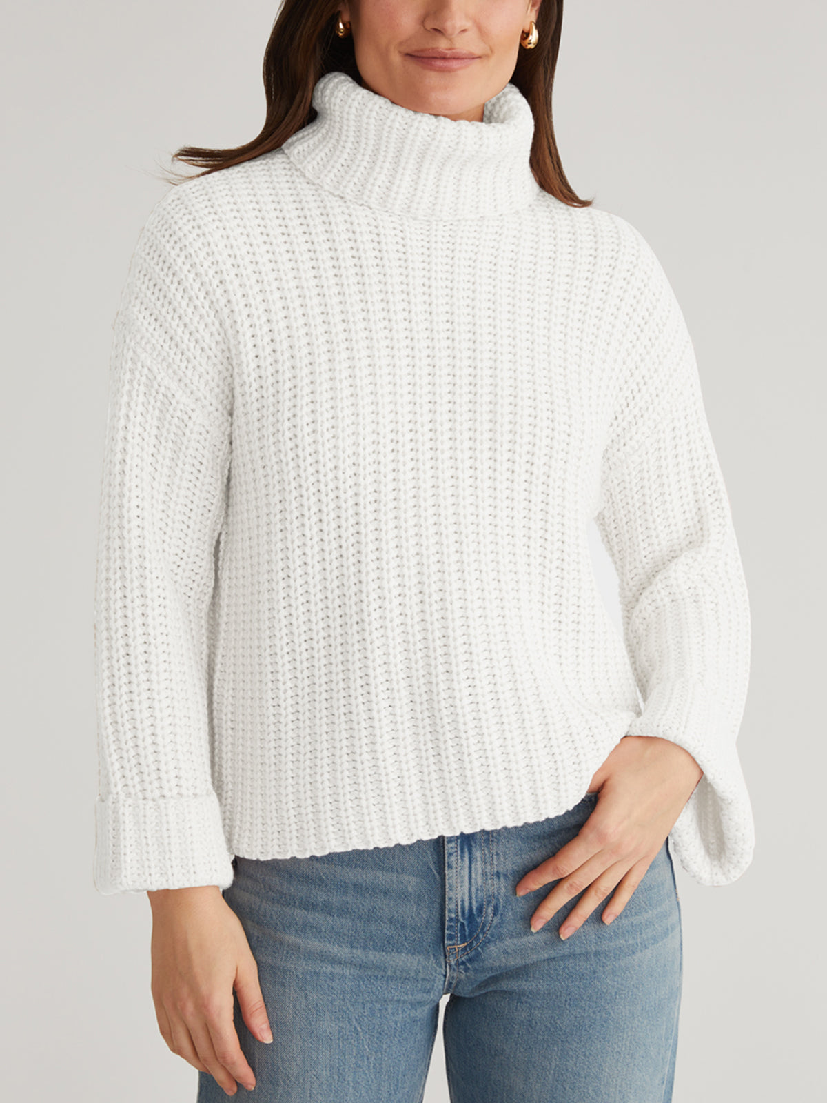 A person stands against a plain background wearing a cozy 525 America white Turtleneck Shaker Pullover with oversized sleeves and blue jeans. The turtleneck frames their face, partially out of frame, as one hand rests on their hip.
