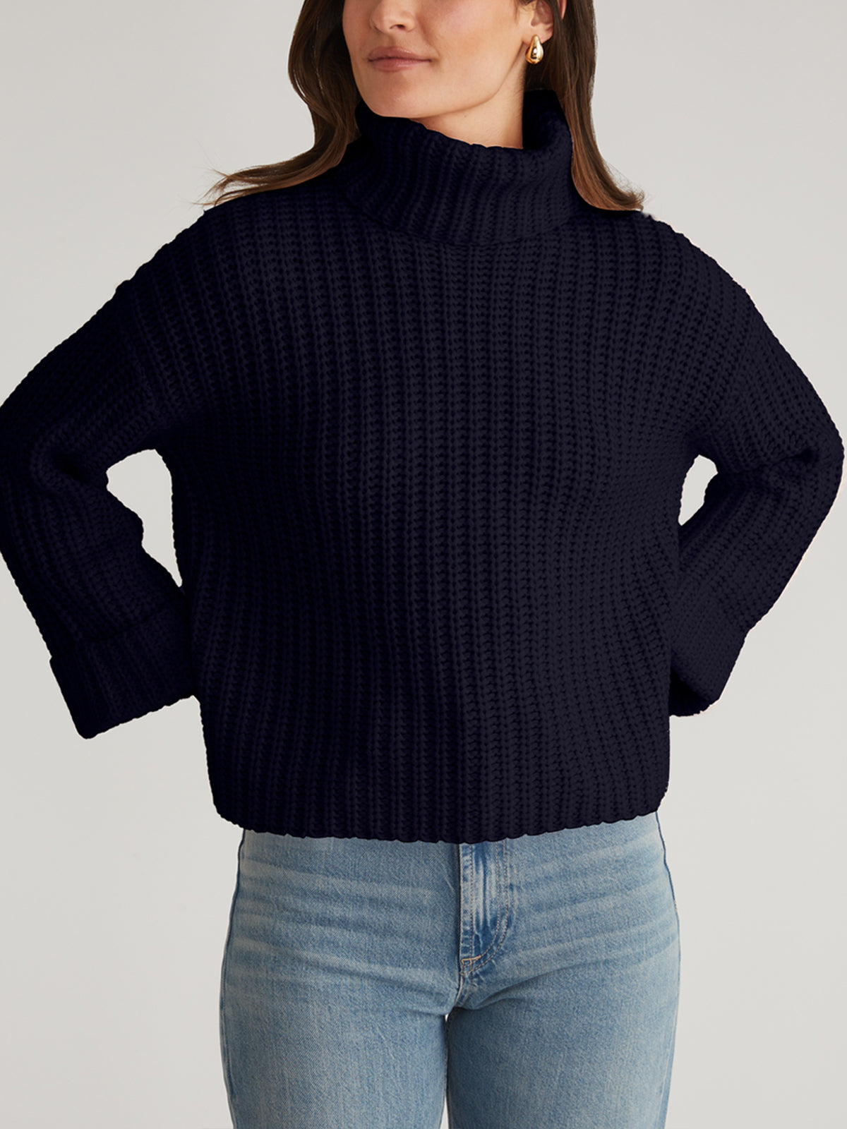 A person stands confidently with hands on hips wearing a chunky, dark blue Turtleneck Shaker Pullover by 525 America. The pullover features oversized sleeves and is paired effortlessly with light blue jeans against a neutral background.