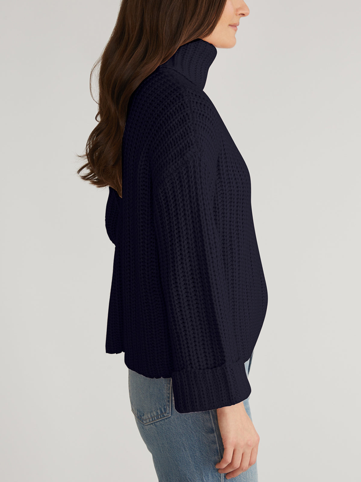 A person is seen from the side in a cozy turtleneck design by 525 America, featuring a textured knit pattern. They pair the dark-colored Turtleneck Shaker Pullover with blue jeans, their long, wavy hair flowing gracefully against a plain backdrop.