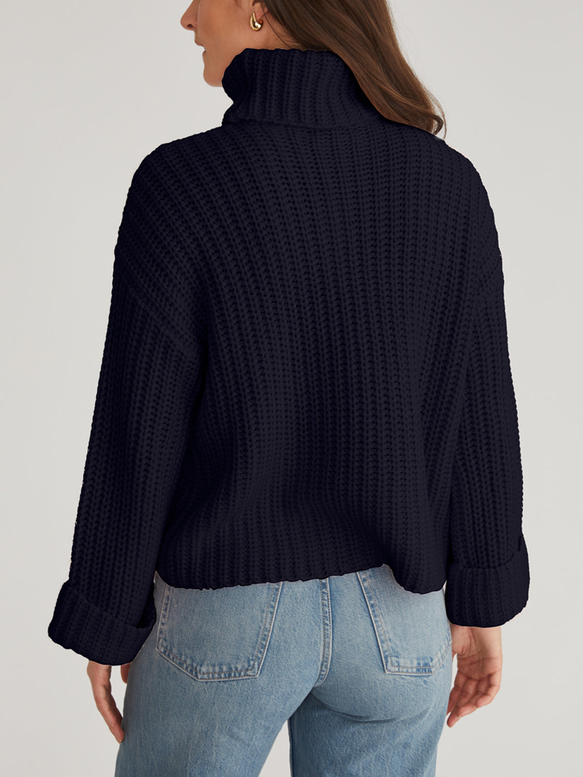 Seen from behind, a person with long hair wears a dark Turtleneck Shaker Pullover by 525 America. The sweater features chunky texture and oversized sleeves, paired with light blue jeans. They face left against a light-colored, plain background.