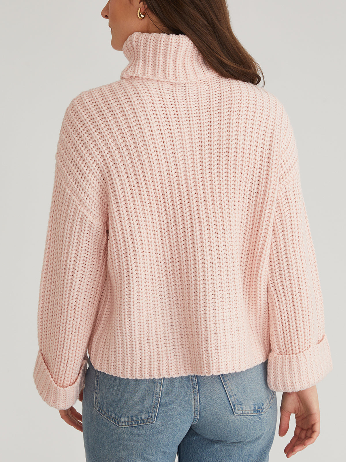The image features a person from behind wearing a chunky, pink Turtleneck Shaker Pullover by 525 America, highlighting its cozy turtleneck and oversized sleeves with cuffs. Its perfectly paired with light blue jeans against a plain, light-colored background.