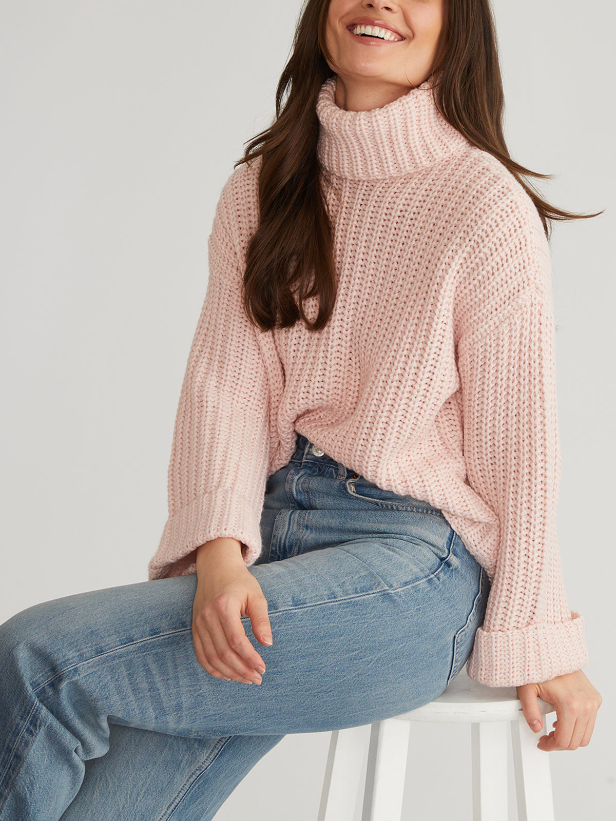 A woman with long brown hair, smiling and wearing blue jeans, sits on a stool in the cozy 525 America Turtleneck Shaker Pullover sweater with oversized sleeves. The image is cropped at her nose, highlighting the stylish design.