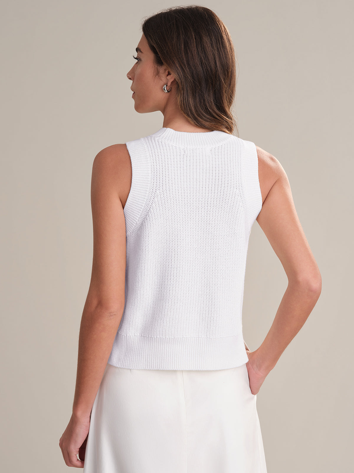 A woman with medium-length brown hair is shown from the back, wearing a 525 America 100% cotton crew neck sleeveless knit top and a white skirt, standing against a plain, light-colored background.