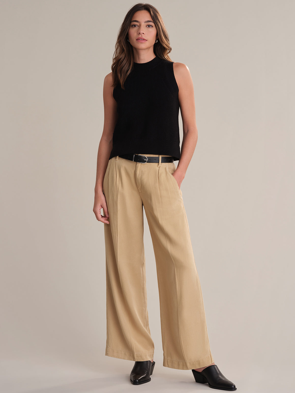 A person poses against a plain background wearing a 525 America black sleeveless 100% cotton top, beige wide-leg trousers with a black belt, and black shoes. Hands in pockets, they look at the camera, embodying effortless layering style.