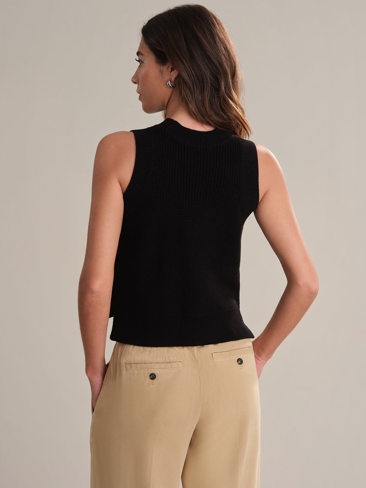 A person with long brown hair wears a sleeveless black knit top from 525 America—ideal for layering—paired with beige trousers, standing with their back to the camera against a plain, light-colored background.