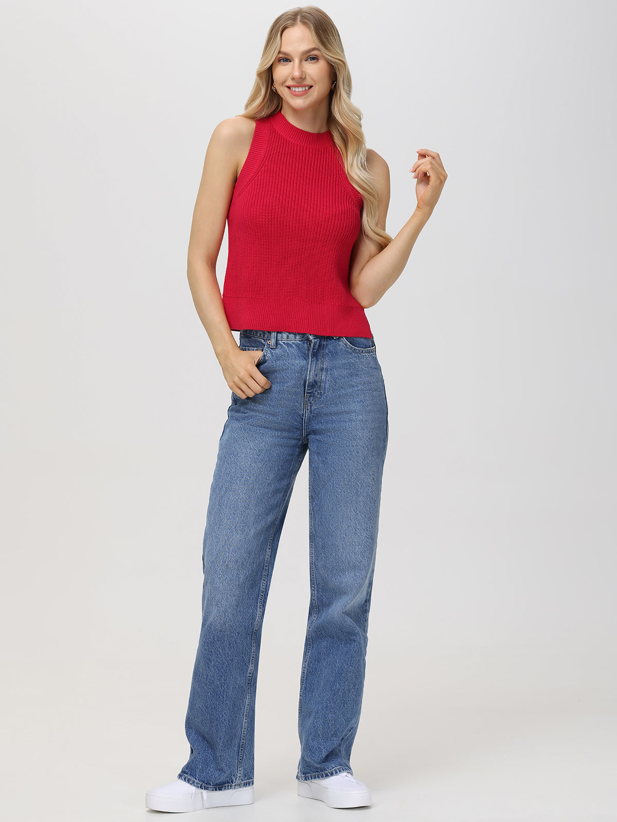 A woman poses against a plain light background, wearing a chic sleeveless red ribbed top by 525 America, paired with blue high-waisted jeans and white sneakers. Her long, wavy blonde hair frames her smile as she raises one hand near her face.