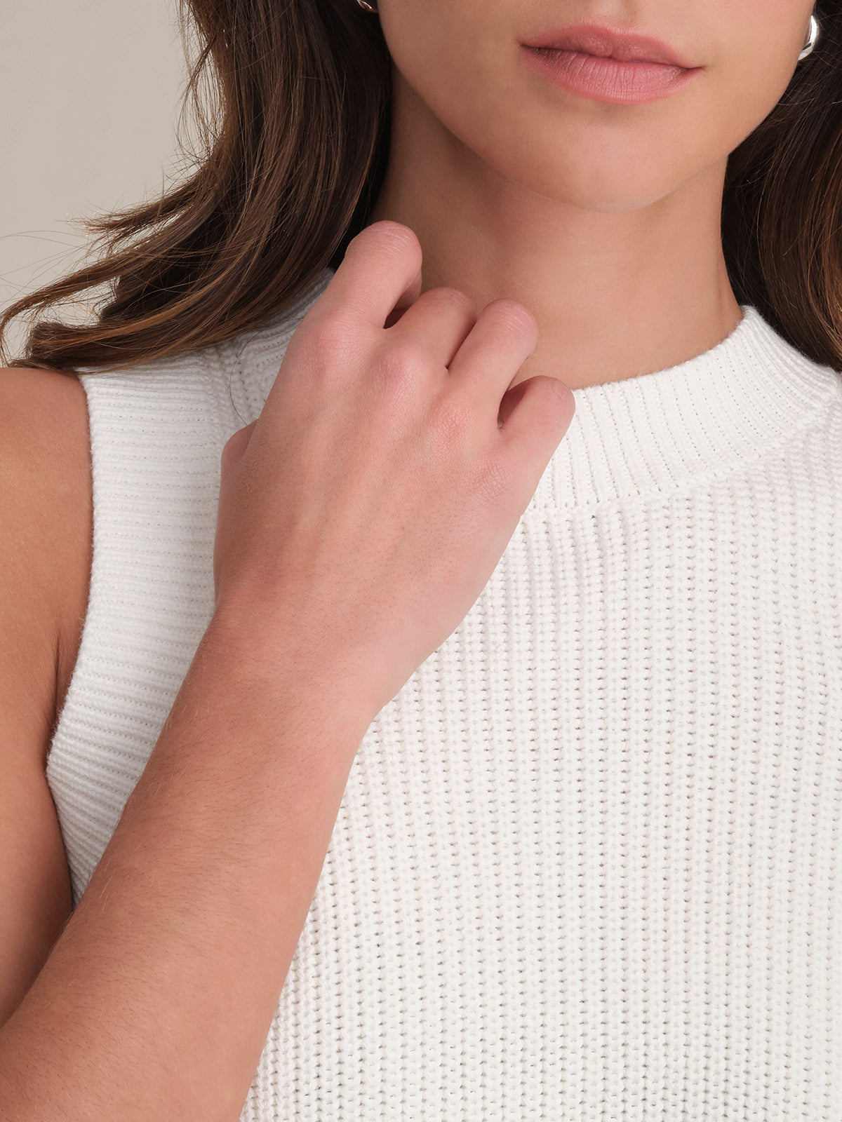 A woman is shown from the shoulders up in a sleeveless, 100% cotton ribbed white top by 525 America. Her right hand rests near her collarbone, and her brown hair is partially visible. The crew neck design adds to the casual comfort of the look.