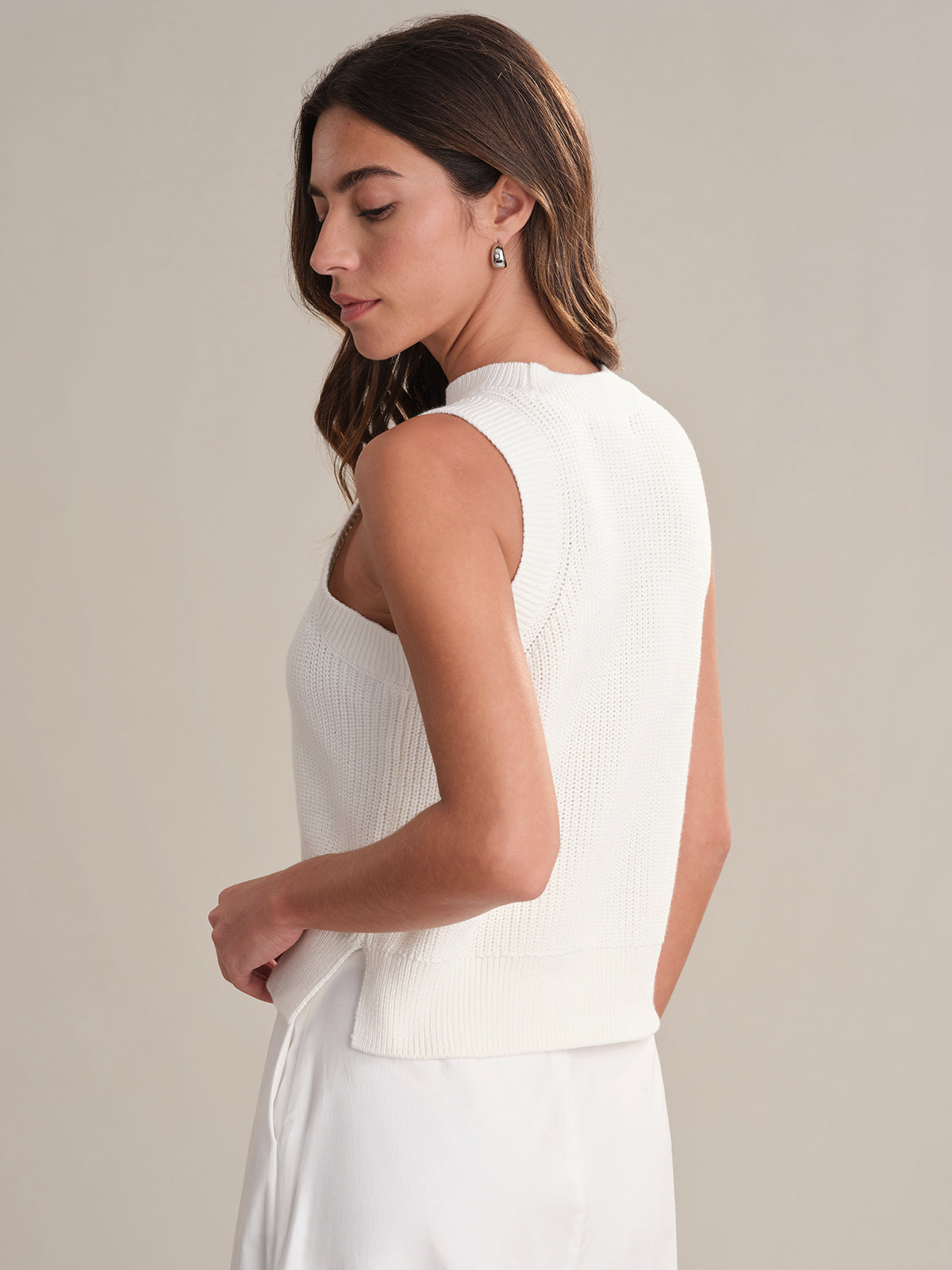 A woman with long brown hair wears a 525 America sleeveless crew neck white knit top and matching white skirt, standing sideways and looking down against a neutral background.