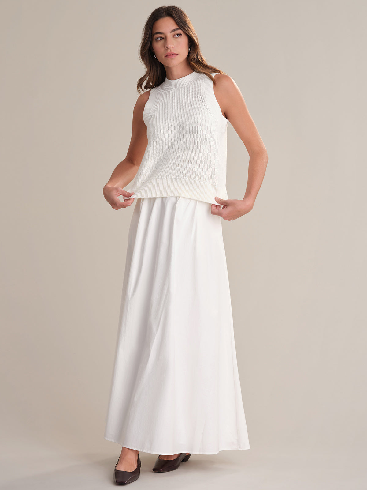 A woman wears a sleeveless white 100% cotton knit crew neck top by 525 America and a long white skirt, standing before a plain background. She holds the sides of her top, embracing casual comfort, and wears dark brown shoes.
