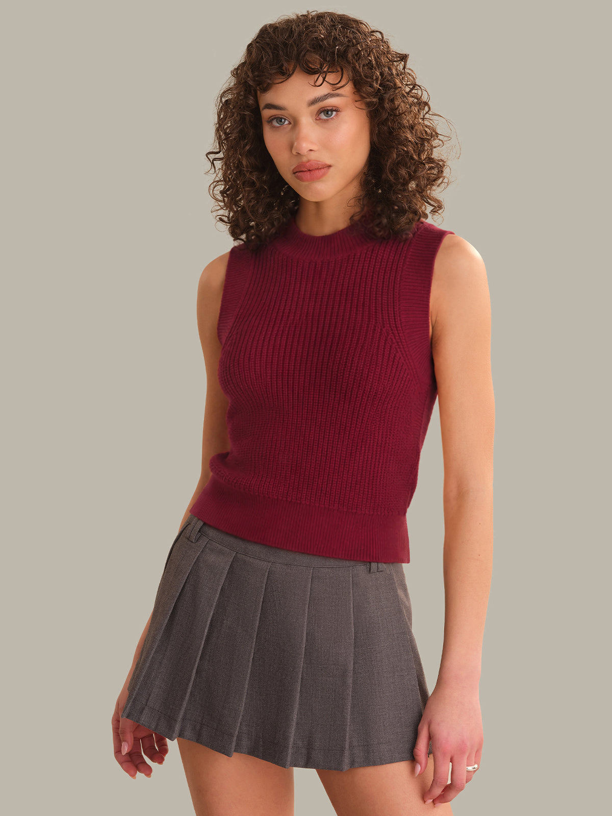 Woman wearing a maroon sleeveless top and gray pleated skirt on a neutral background