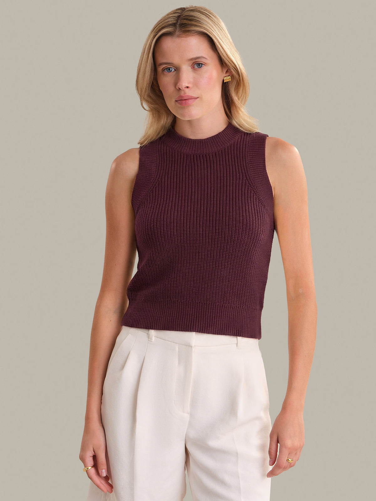 Woman wearing a sleeveless burgundy top and white pants against a neutral background