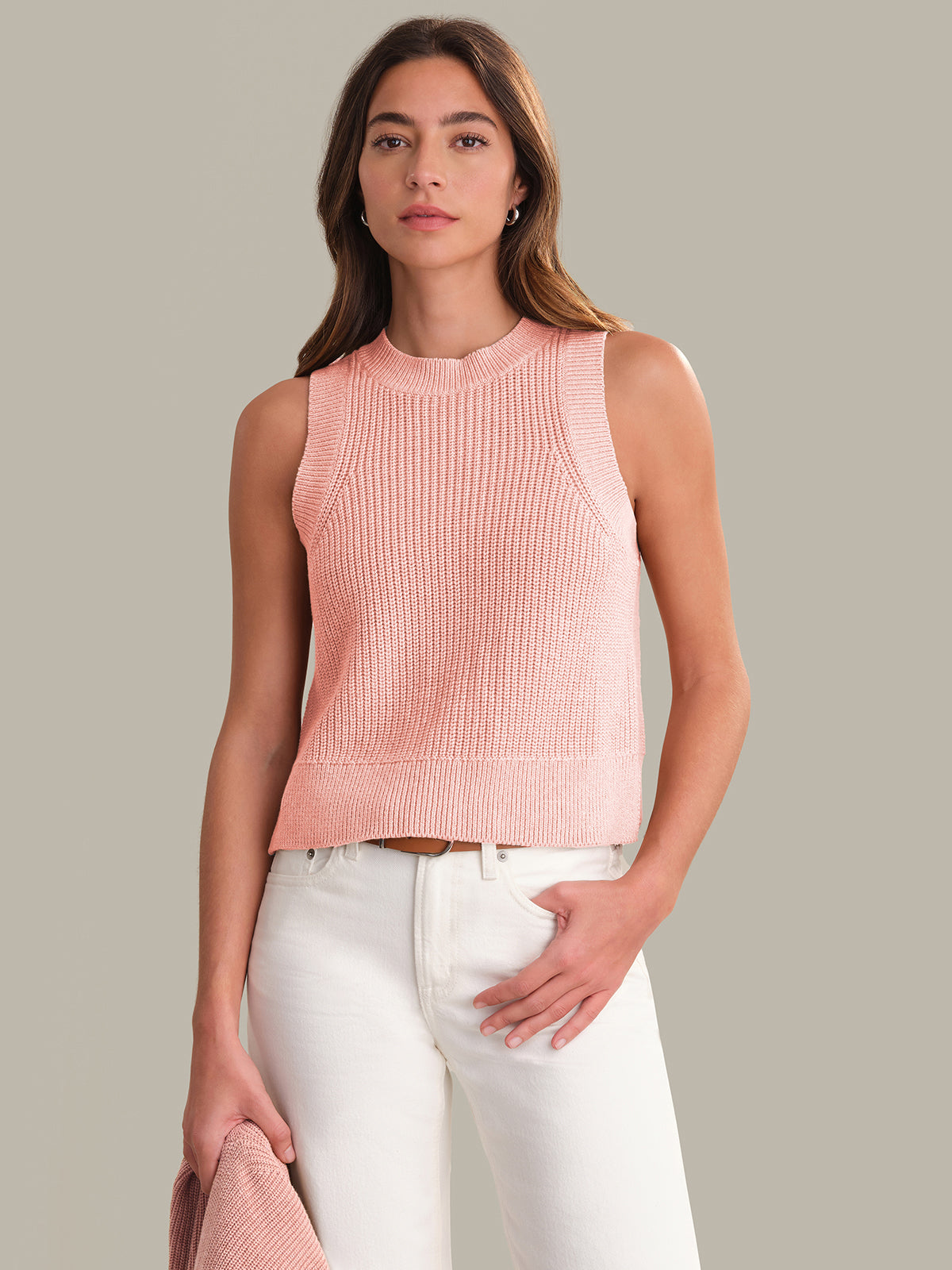 Woman wearing a pink sleeveless knitted top and white pants against a neutral background