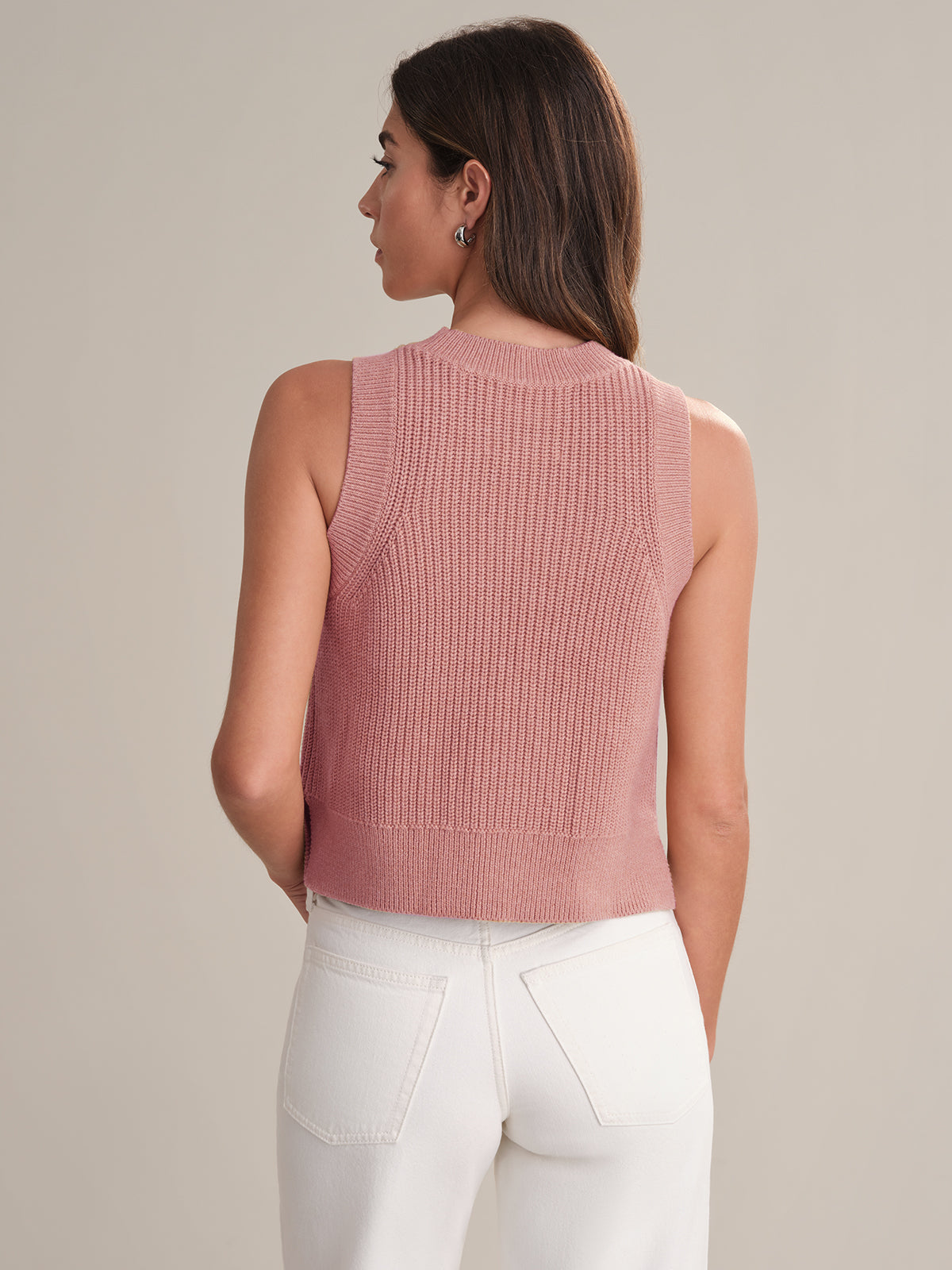 A woman with long brown hair is seen from behind wearing a sleeveless light pink knit top from 525 America and high-waisted white pants, creating an effortlessly chic, versatile look against a neutral background.