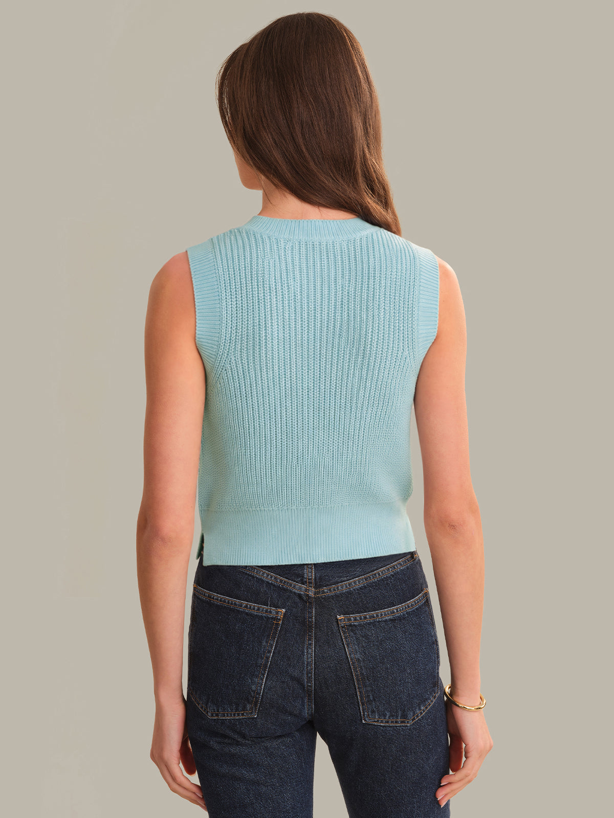 Woman wearing a light blue sleeveless sweater and dark jeans against a plain background