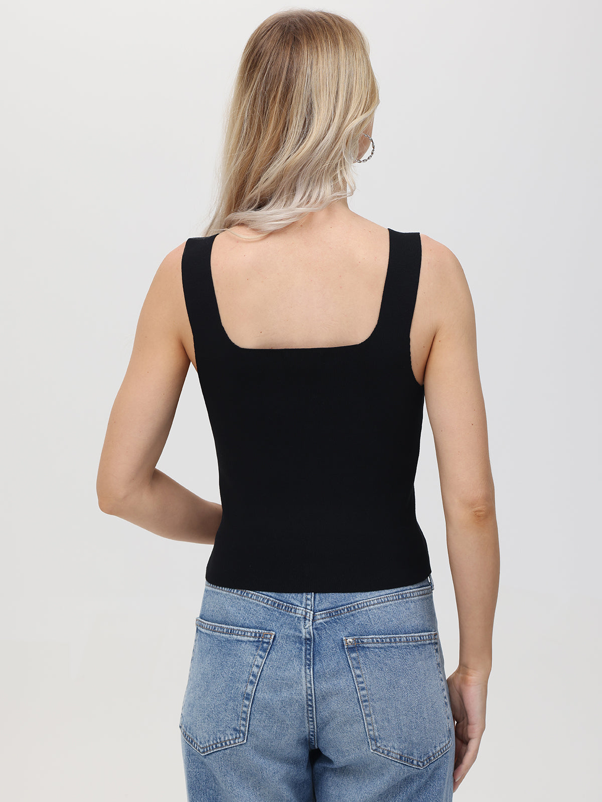 A person with long blonde hair, turned away from the camera, wears a black sleeveless top and blue form-fitting jeans by 525 America. The plain white background showcases their styles versatility, making it perfect for layering on any occasion.