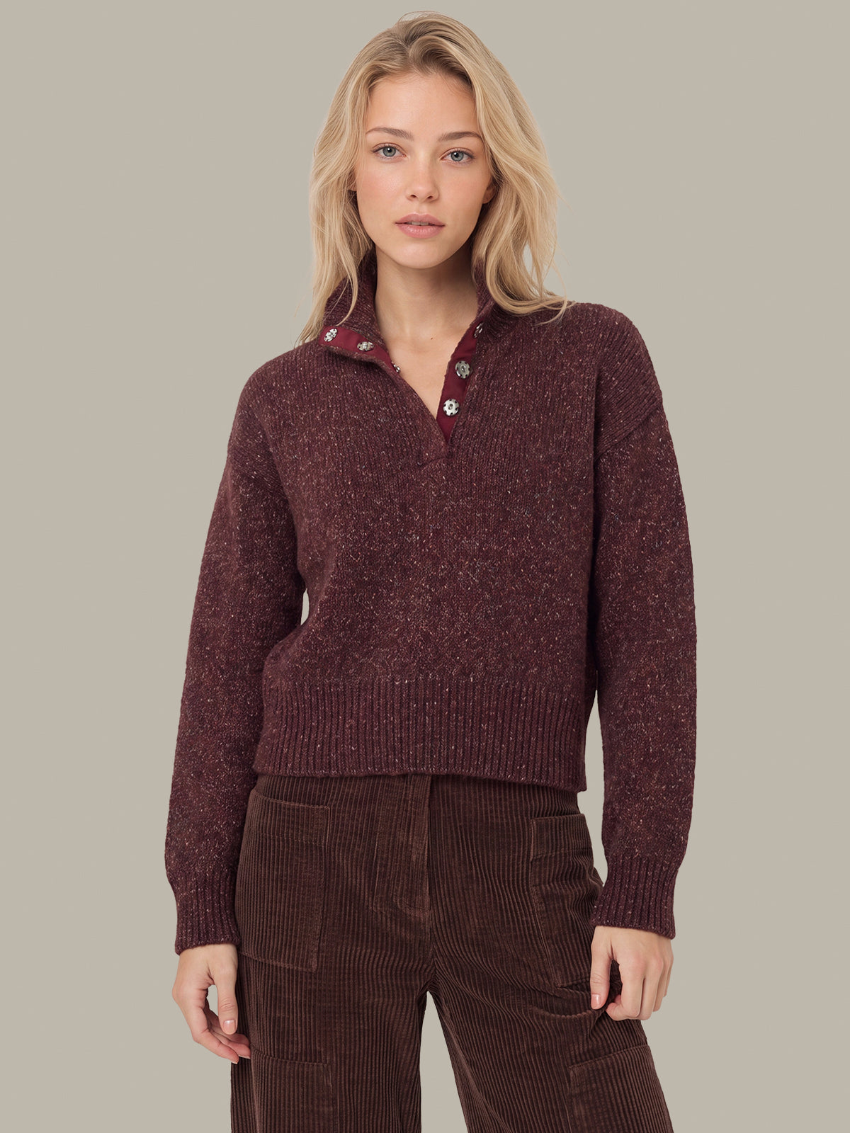 A woman modeling a marled burgundy knit half-placket sweater with a ribbed collar and brown corduroy pants, set against a solid warm gray studio background.