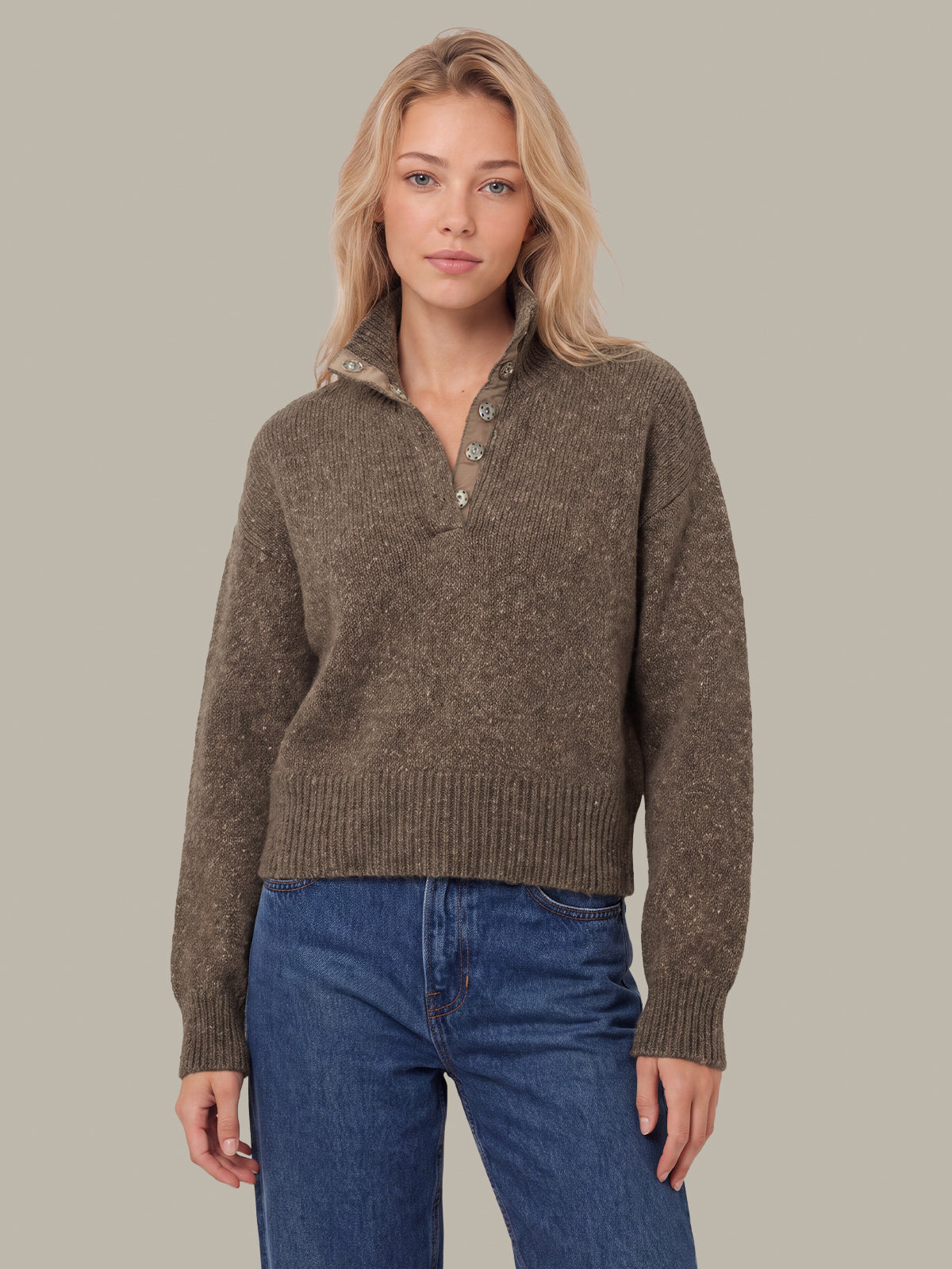 A woman modeling a brown textured knit half-placket sweater with a buttoned collar, paired with blue jeans, against a solid beige background.