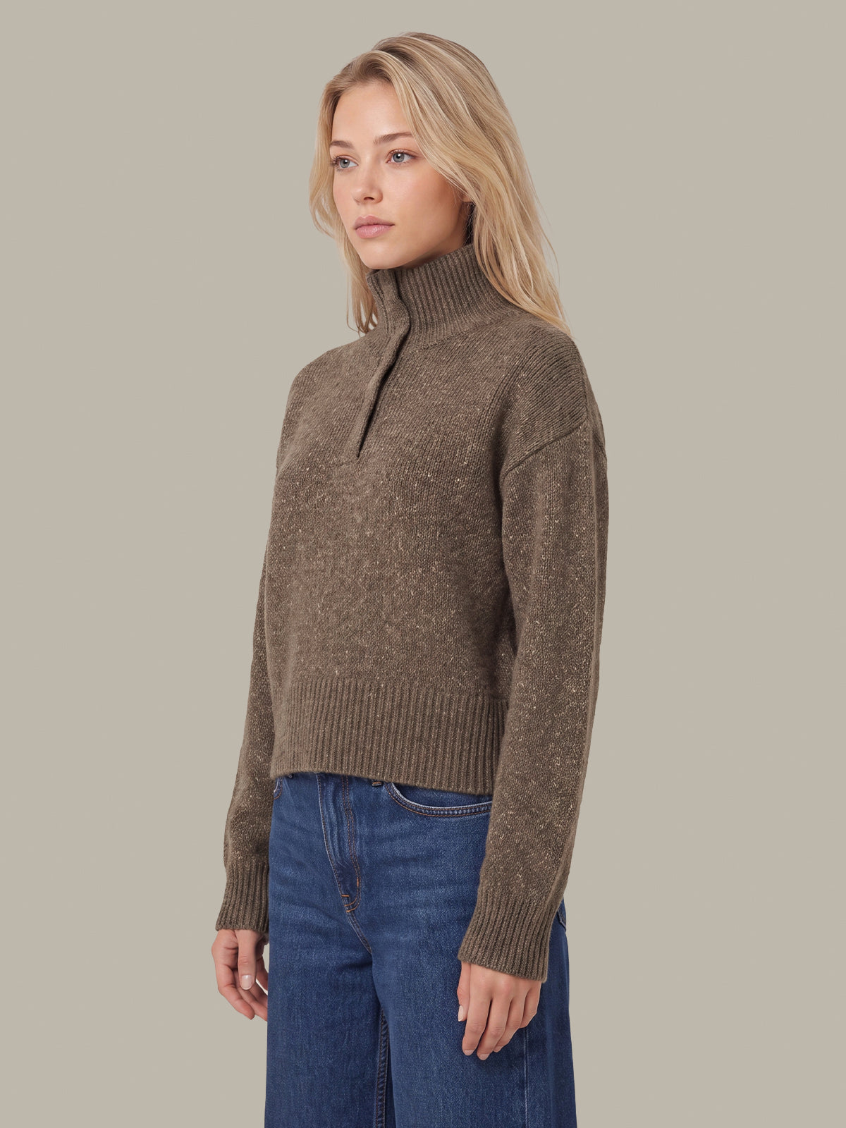 A woman modeling a brown ribbed knit mock neck sweater with a short zipper placket, paired with blue jeans, against a solid beige background.