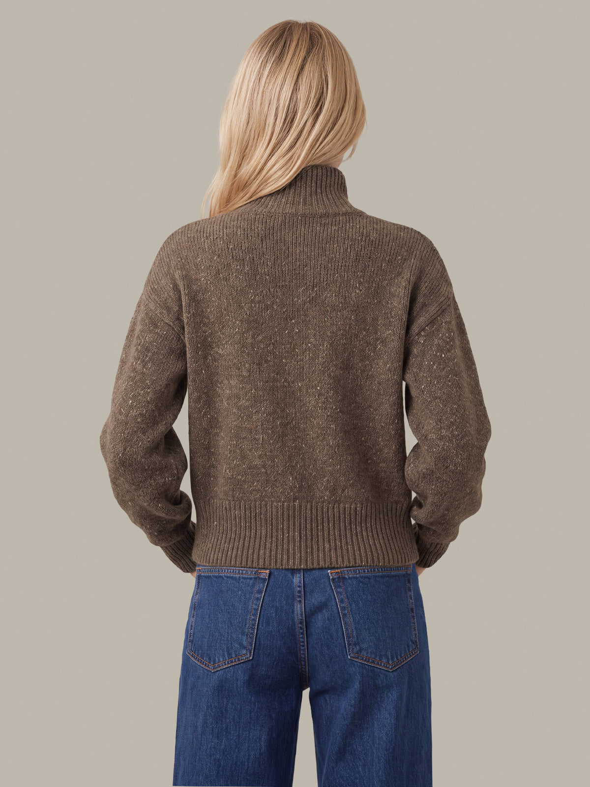A woman viewed from the back wearing a brown ribbed knit mock-neck sweater and blue jeans, set against a solid light gray background.