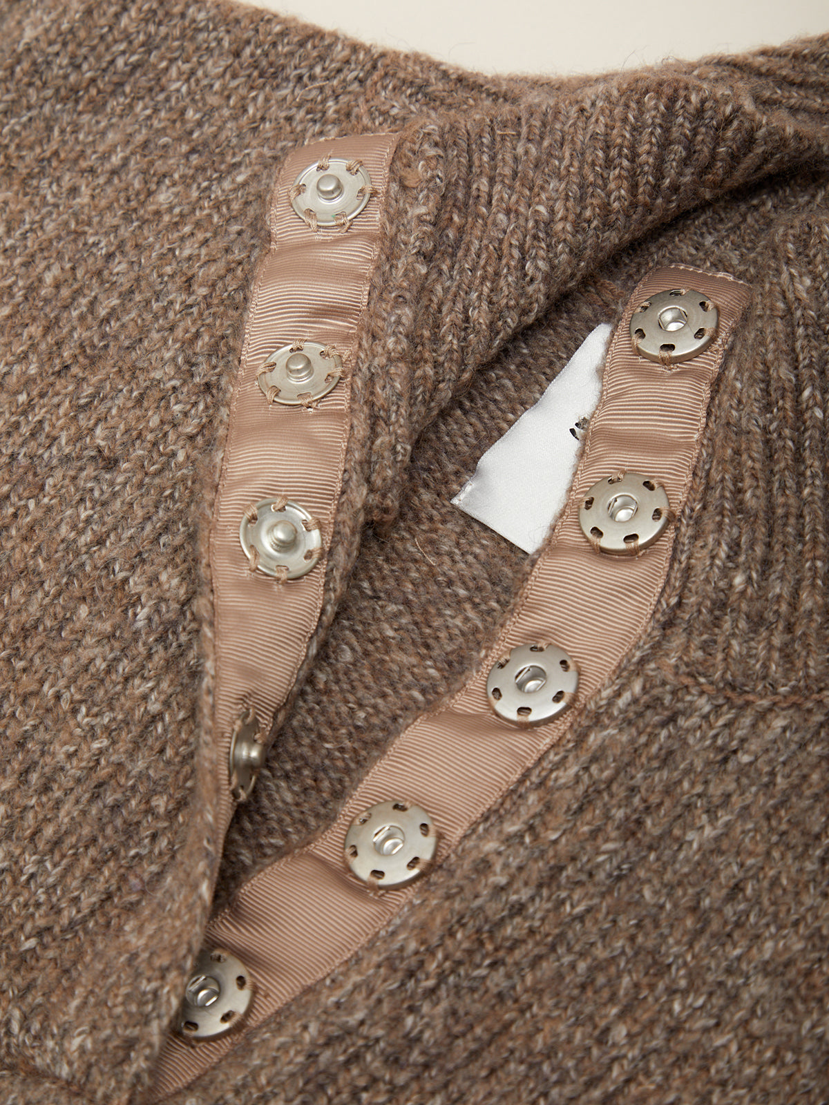 Close-up of a brown knit sweater with a ribbed trim and decorative silver-tone buttons, showing part of the interior collar and a white brand tag.