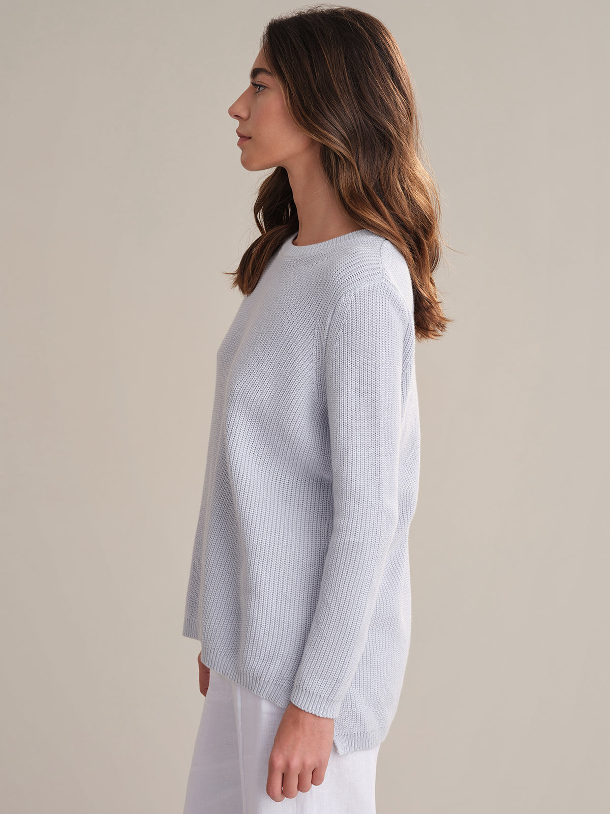 A woman with long, wavy brown hair stands in profile against a plain background, wearing a 525 America relaxed-fit, 100% cotton ribbed long-sleeve sweater and white pants.