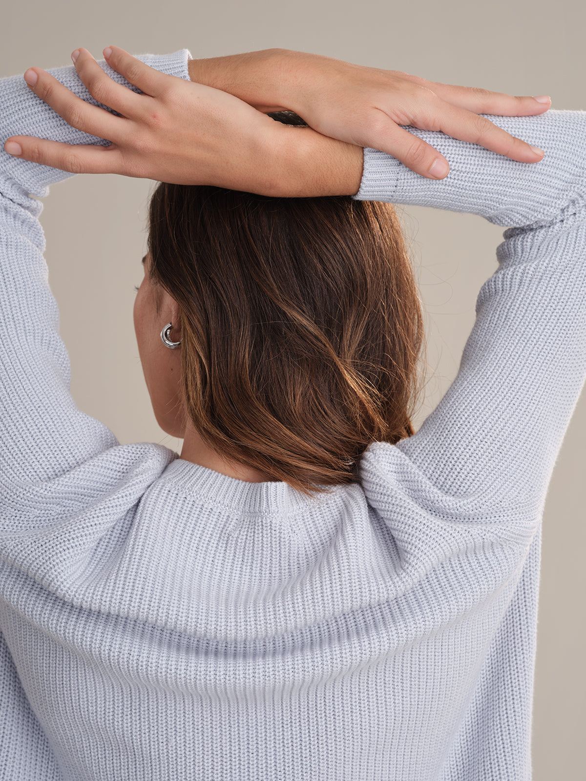 A person with medium-length brown hair, wearing a relaxed fit, 100% cotton light gray knit sweater by 525 America, stands with their back to the camera and arms raised, crossing their forearms above their head against a plain background.