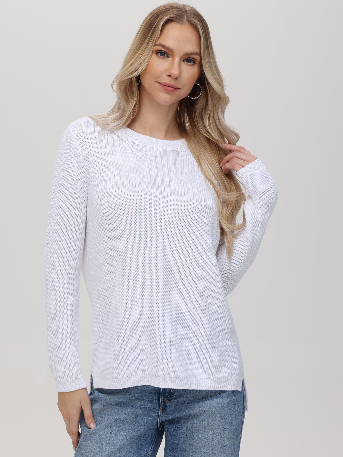 A woman with long, wavy blonde hair wears a relaxed-fit 100% cotton white knit sweater by 525 America and blue jeans. She stands against a plain light background, facing the camera with a slight smile.