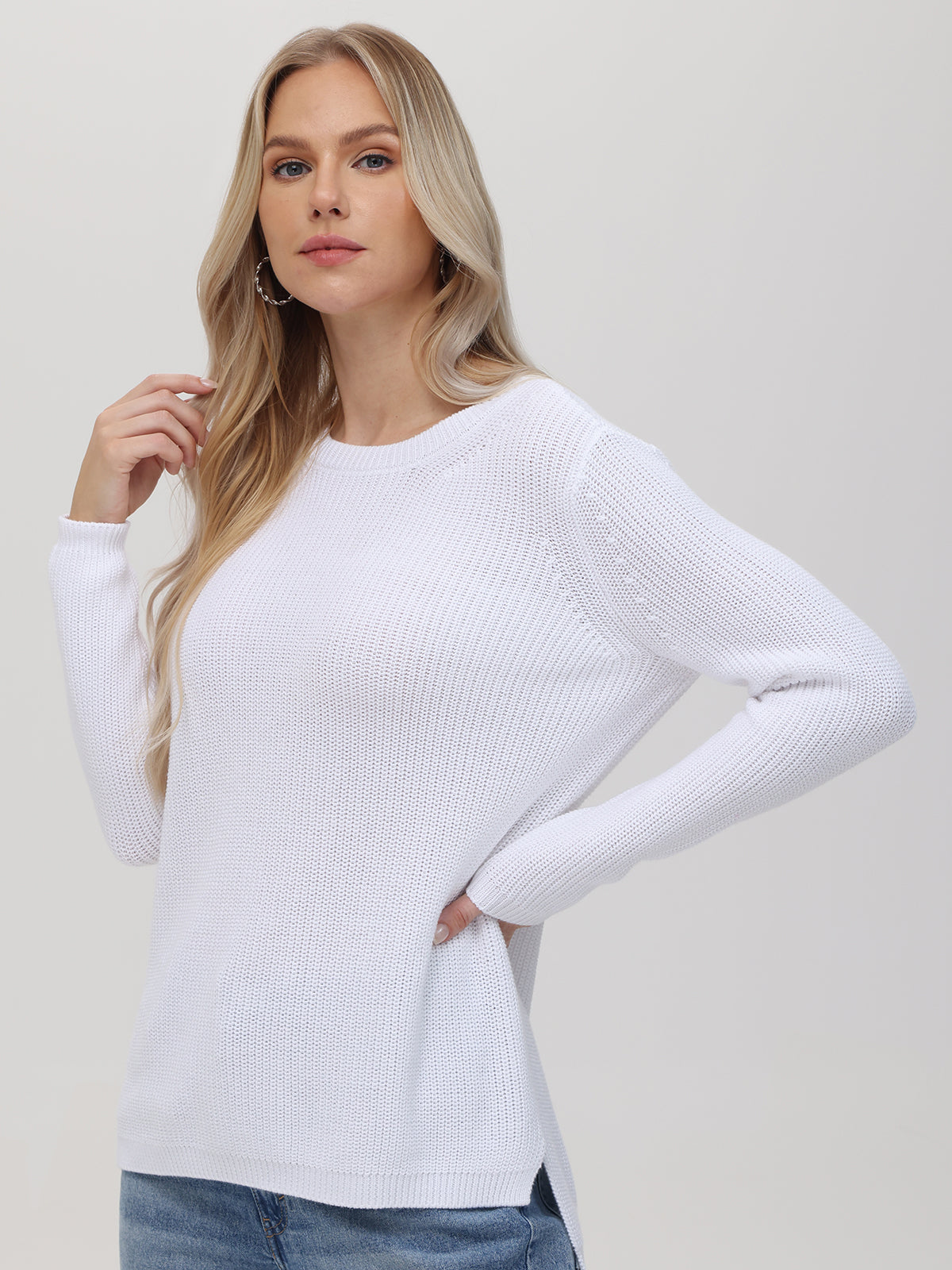 A person with long blonde hair wears a 525 America white, textured shaker stitch knit sweater in a relaxed fit and blue jeans, standing against a plain light background, looking forward with one hand raised near their face.
