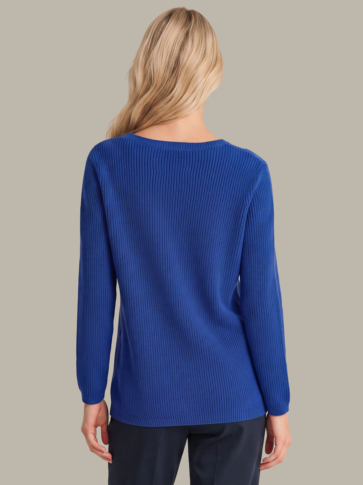 A woman viewed from the back wearing a royal blue ribbed knit sweater and dark pants, set against a solid light gray background.