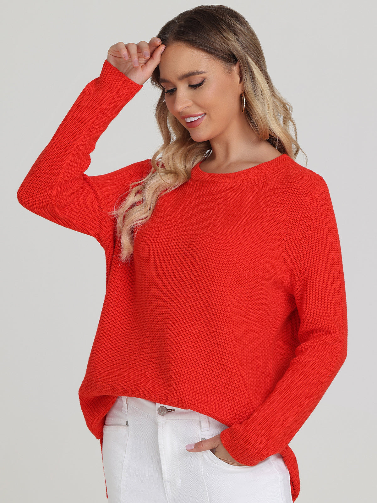 A person with long hair smiles in a bright red shaker stitch sweater by 525 America, highlighting its versatile style. They pair it with white pants, raising one hand near their head while the other rests in their pocket, contrasting beautifully against the plain, light-colored background.