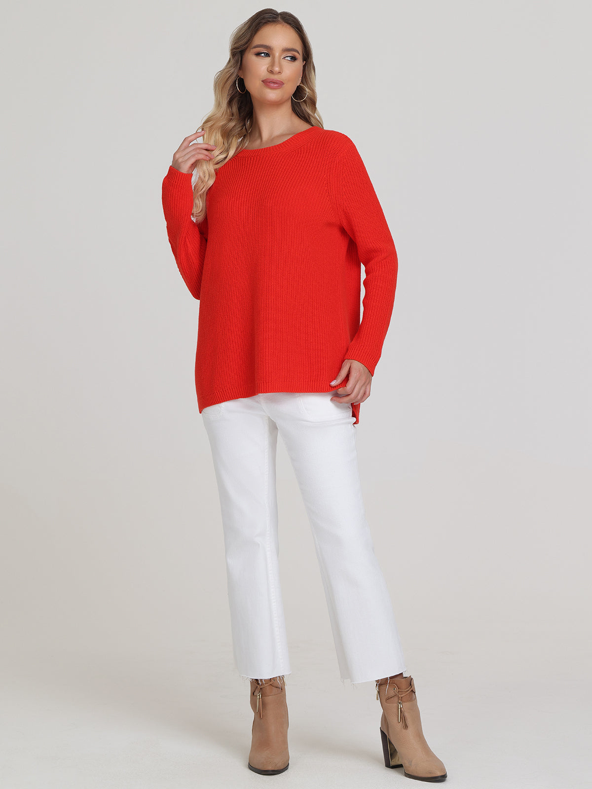 A woman poses against a simple backdrop wearing a bright red shaker stitch sweater by 525 America and white pants. Her long hair falls as she touches it, enhancing her relaxed look, which she completes with brown ankle boots.