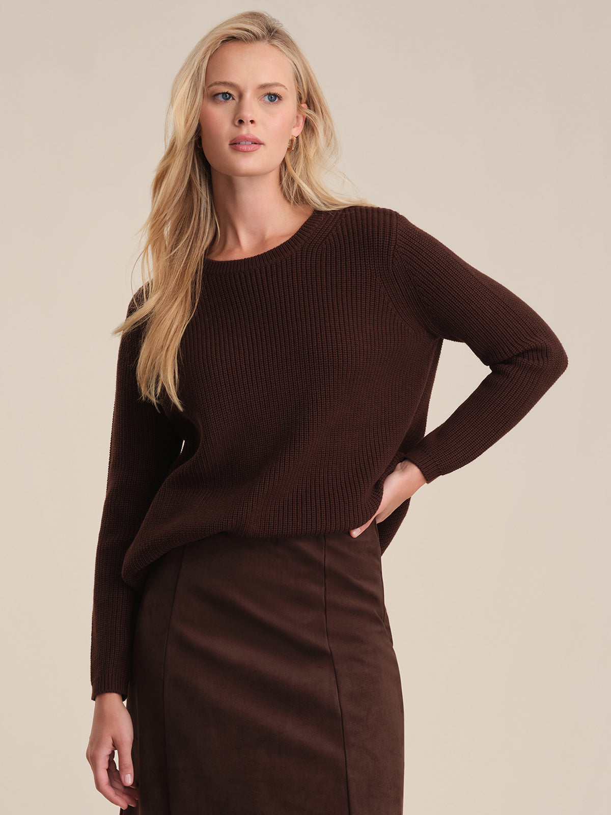 A woman with long blonde hair models a 525 America dark brown ribbed cotton sweater and matching skirt in a relaxed fit, set against a beige background as she gazes sideways with her hand on her hip.