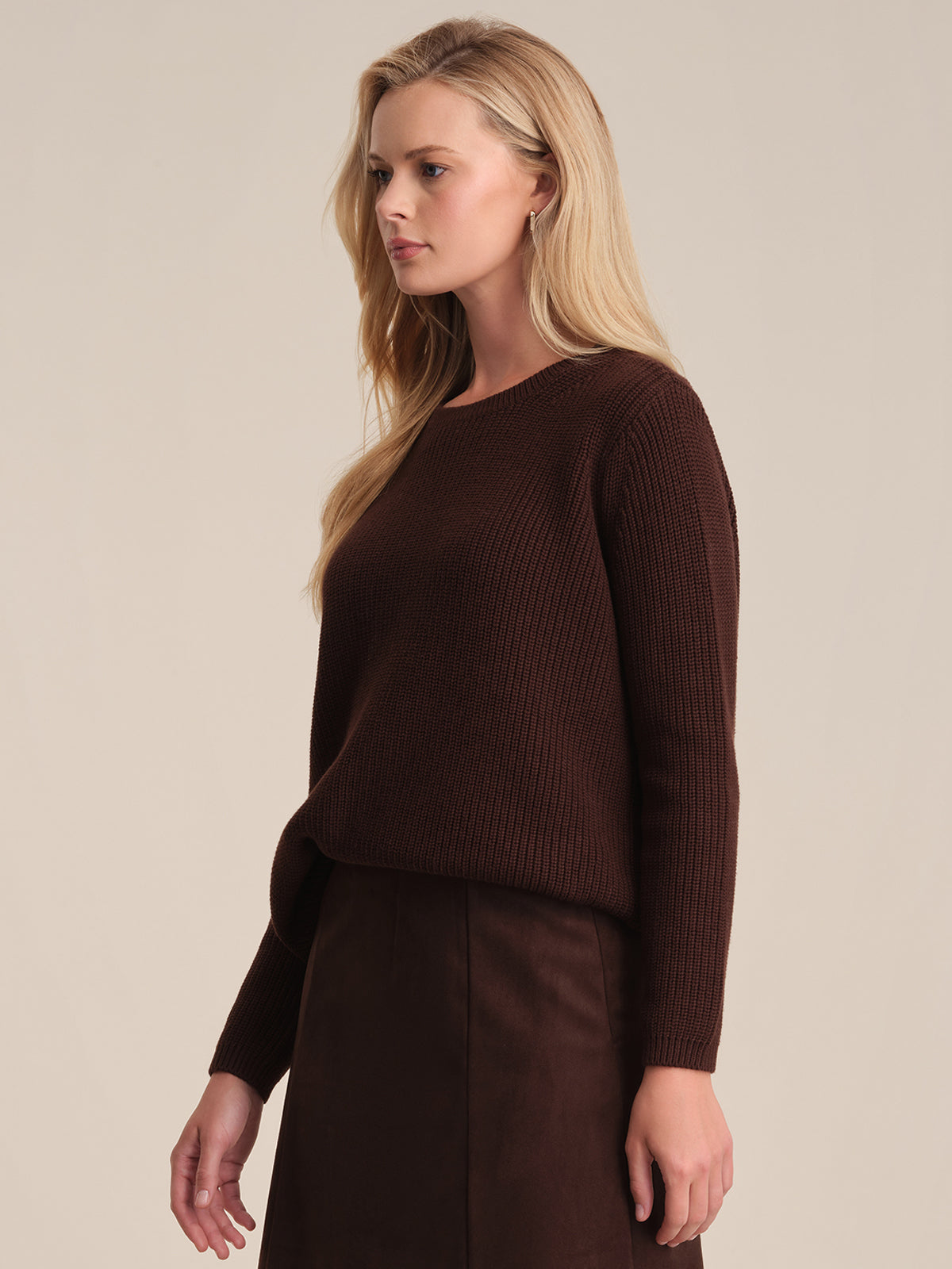 A woman with long blonde hair models a dark brown, relaxed fit shaker stitch sweater and matching skirt by 525 America. Made from 100% cotton, her versatile outfit stands out as she faces slightly left against a plain beige background.