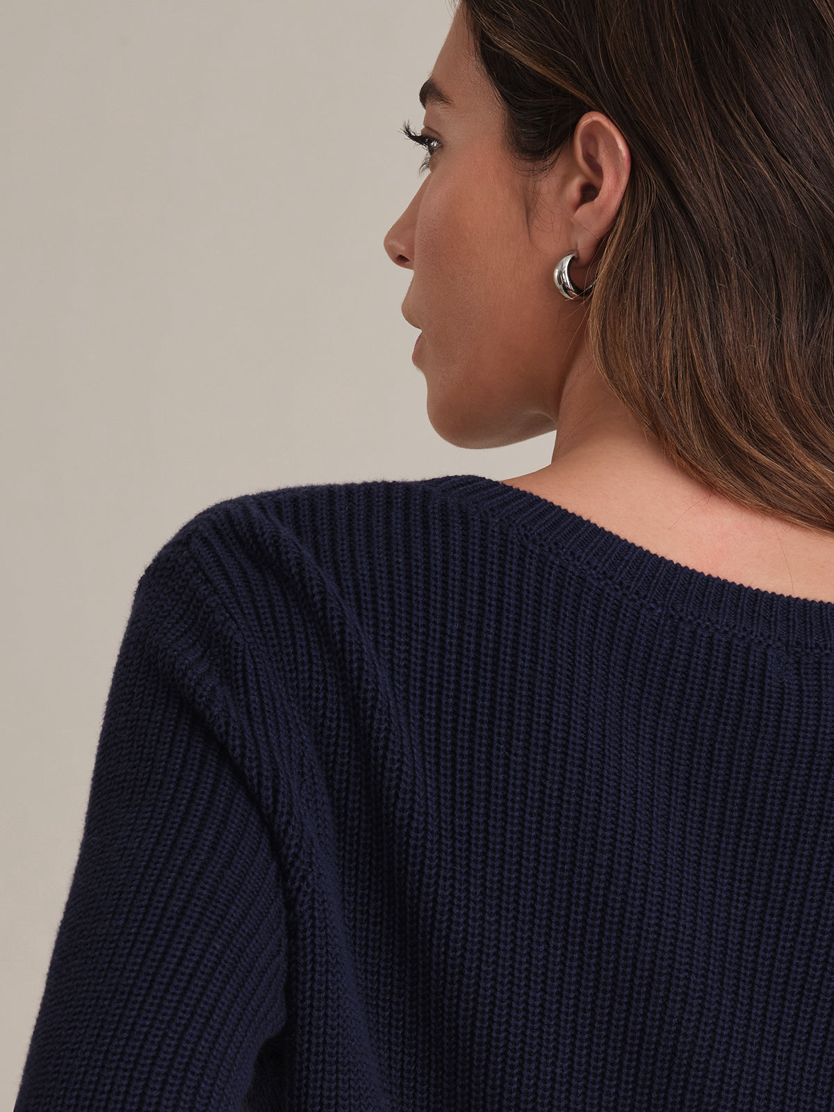 A woman, seen from behind in profile, wears a 100% cotton, dark blue shaker-stitch sweater with a relaxed fit by 525 America. Her brown hair is side-parted, and she stands before a plain light background.