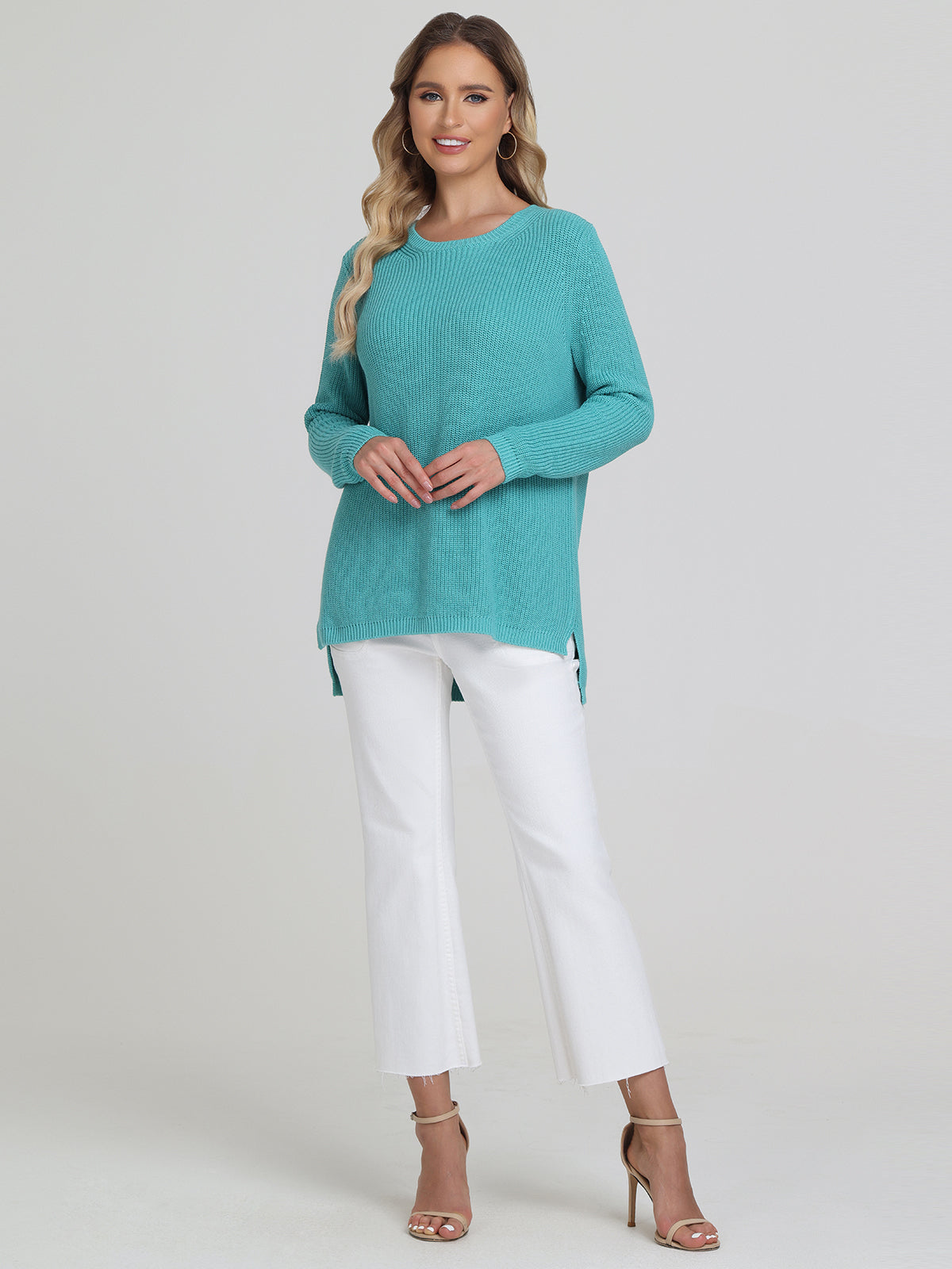 A person models a turquoise 525 America shaker stitch long-sleeved sweater with white cropped pants featuring side slits, paired with beige high-heeled sandals against a plain light gray background, highlighting a versatile style.