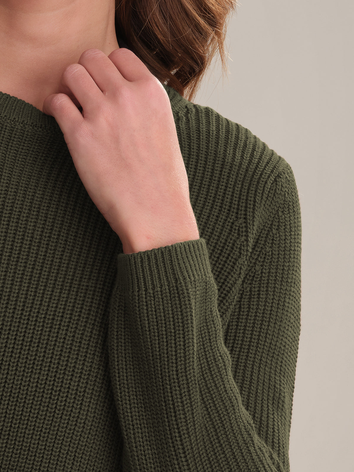 A person in a dark green 525 America knit sweater with a versatile shaker stitch and relaxed fit holds their hand near their collarbone. Their brown hair is partly visible against a plain, neutral background.