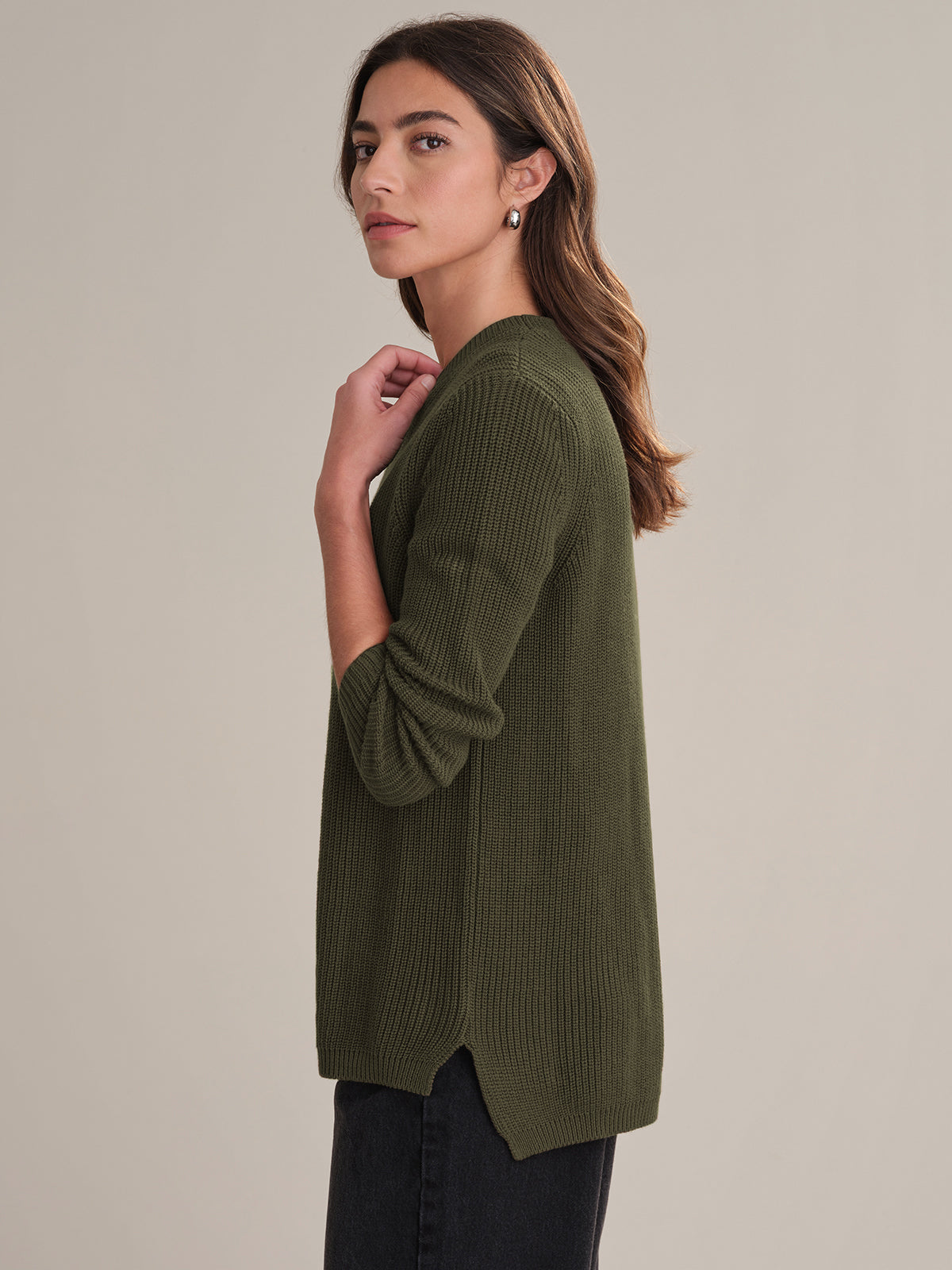 A woman with long brown hair, wearing a 525 America olive green 100% cotton ribbed sweater and black pants, stands sideways and looks at the camera against a plain beige background.