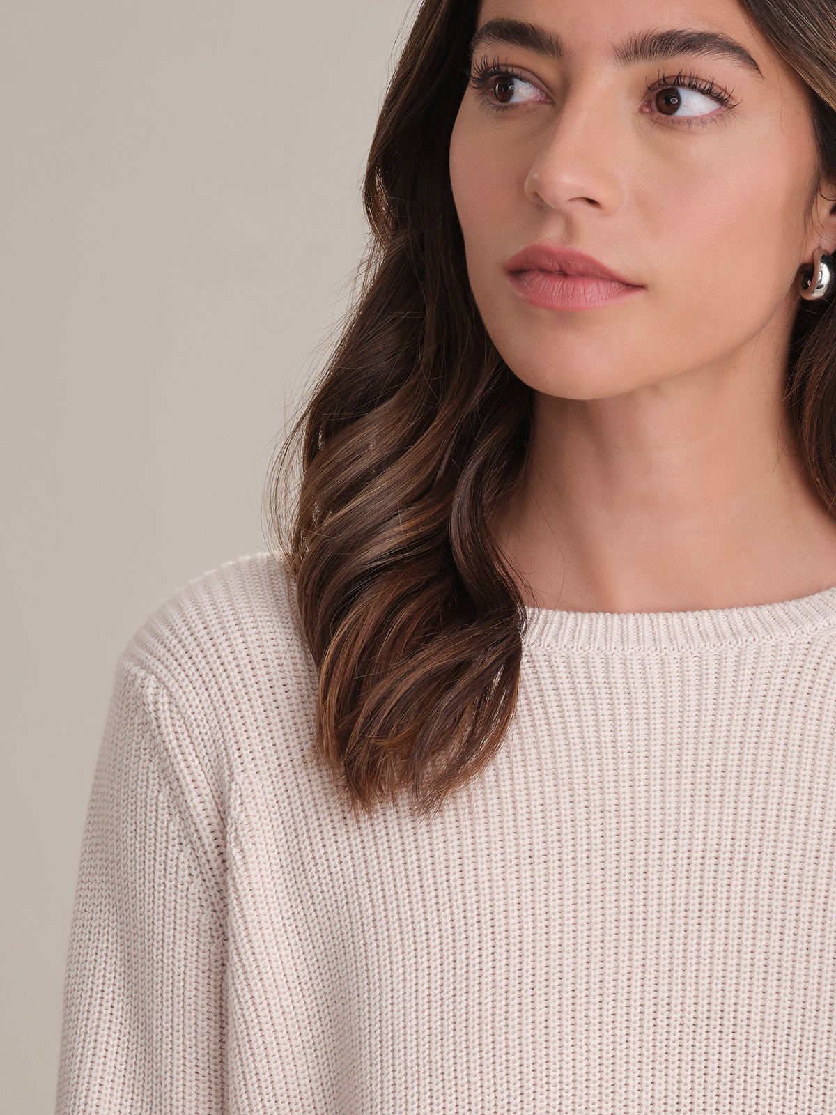 A woman with wavy brown hair and pearl earrings is shown from the shoulders up, looking left. She wears a light beige 525 America shaker stitch sweater made of 100% cotton with a relaxed fit, set against a plain background.