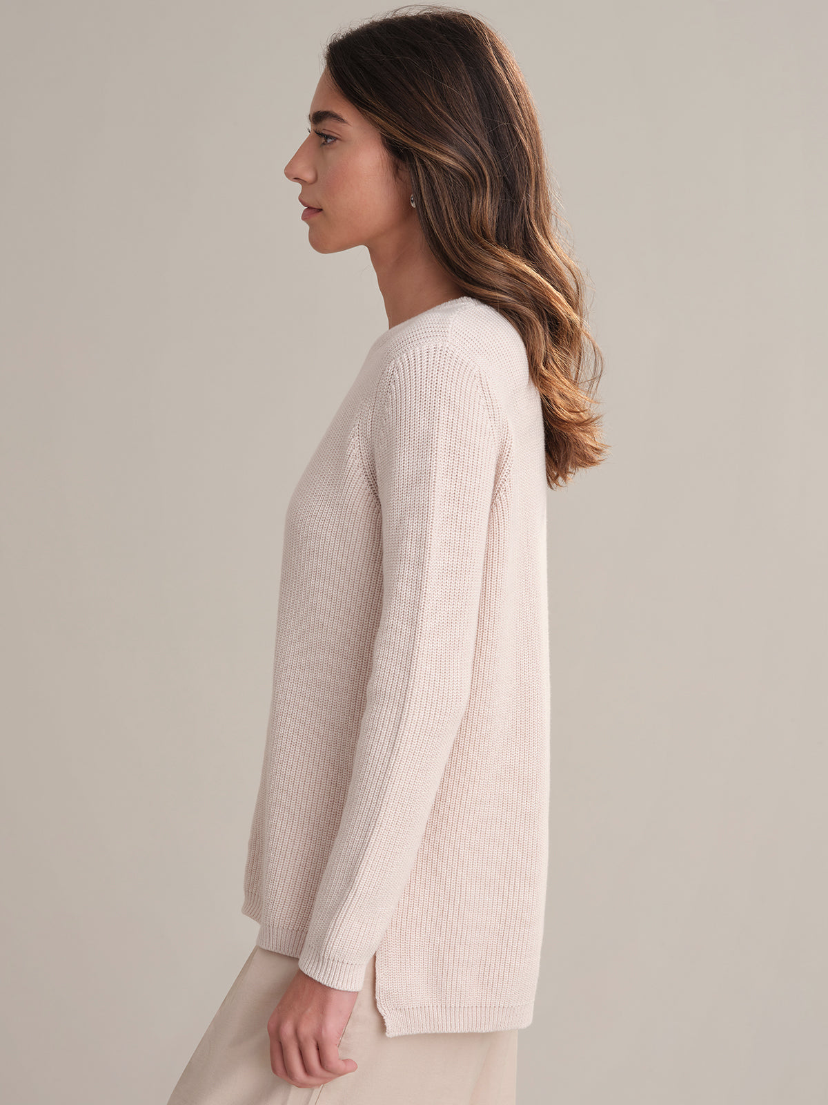 A woman with long brown hair, wearing a relaxed-fit light beige long-sleeve knit sweater from 525 America, stands in profile against a plain light-colored background.