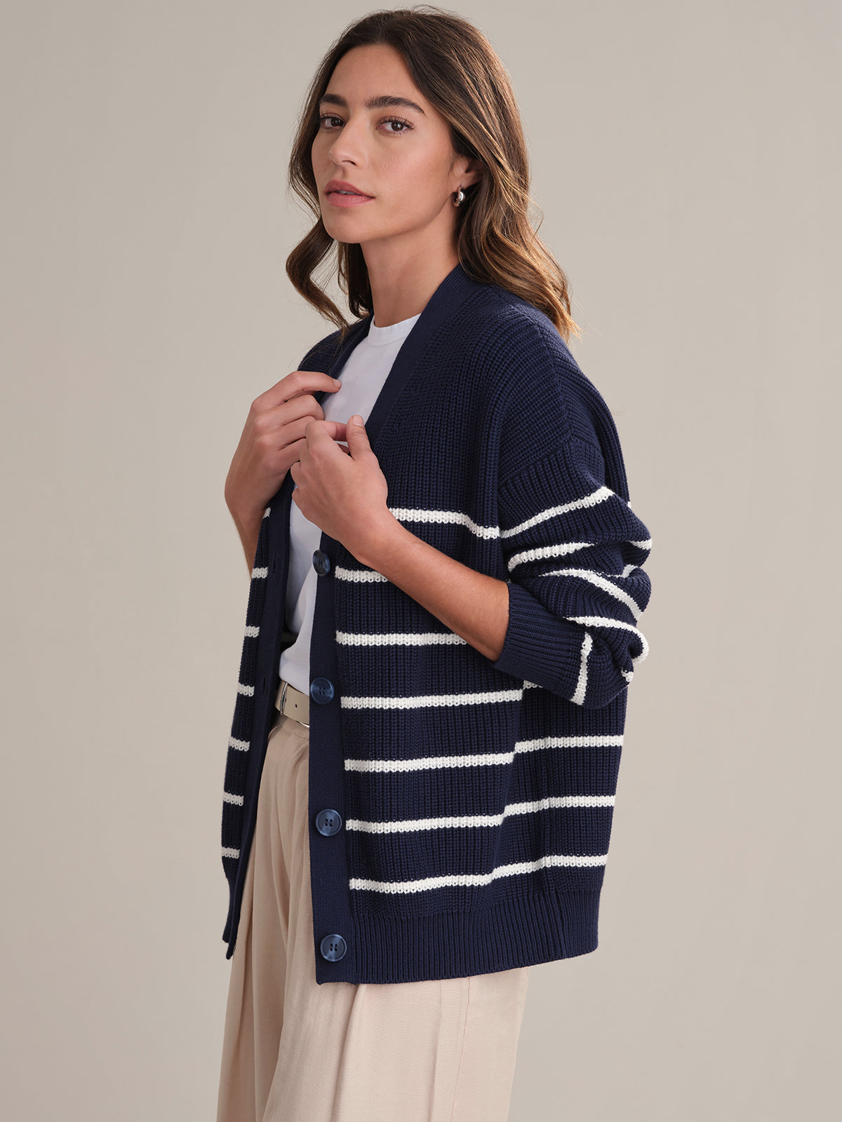 A person with wavy brown hair wears a 525 America relaxed fit navy blue cardigan with white stripes over a white shirt and beige pants, standing against a plain light background.