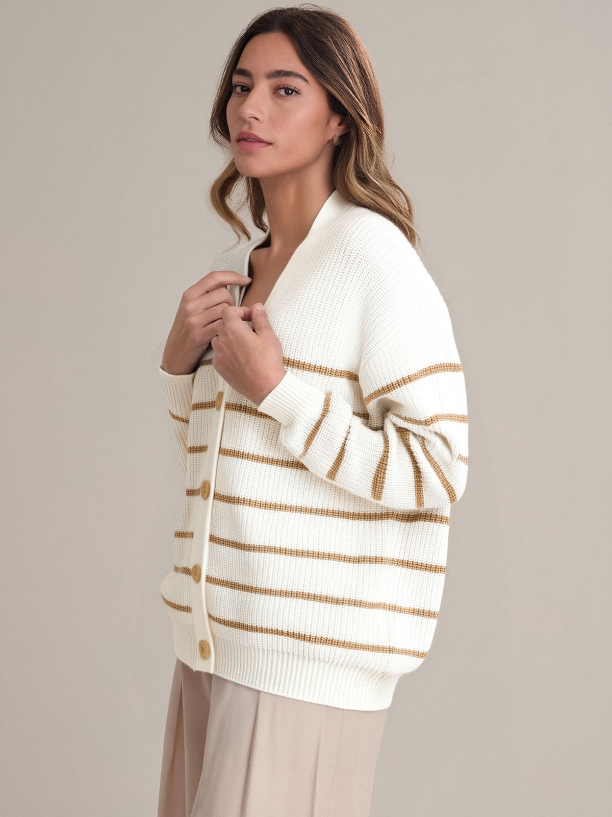 A woman models a 525 America cream cardigan with brown stripes and beige buttons, made from 100% cotton, paired with beige pants. She stands against a plain background, gazing slightly to the side with a neutral expression.