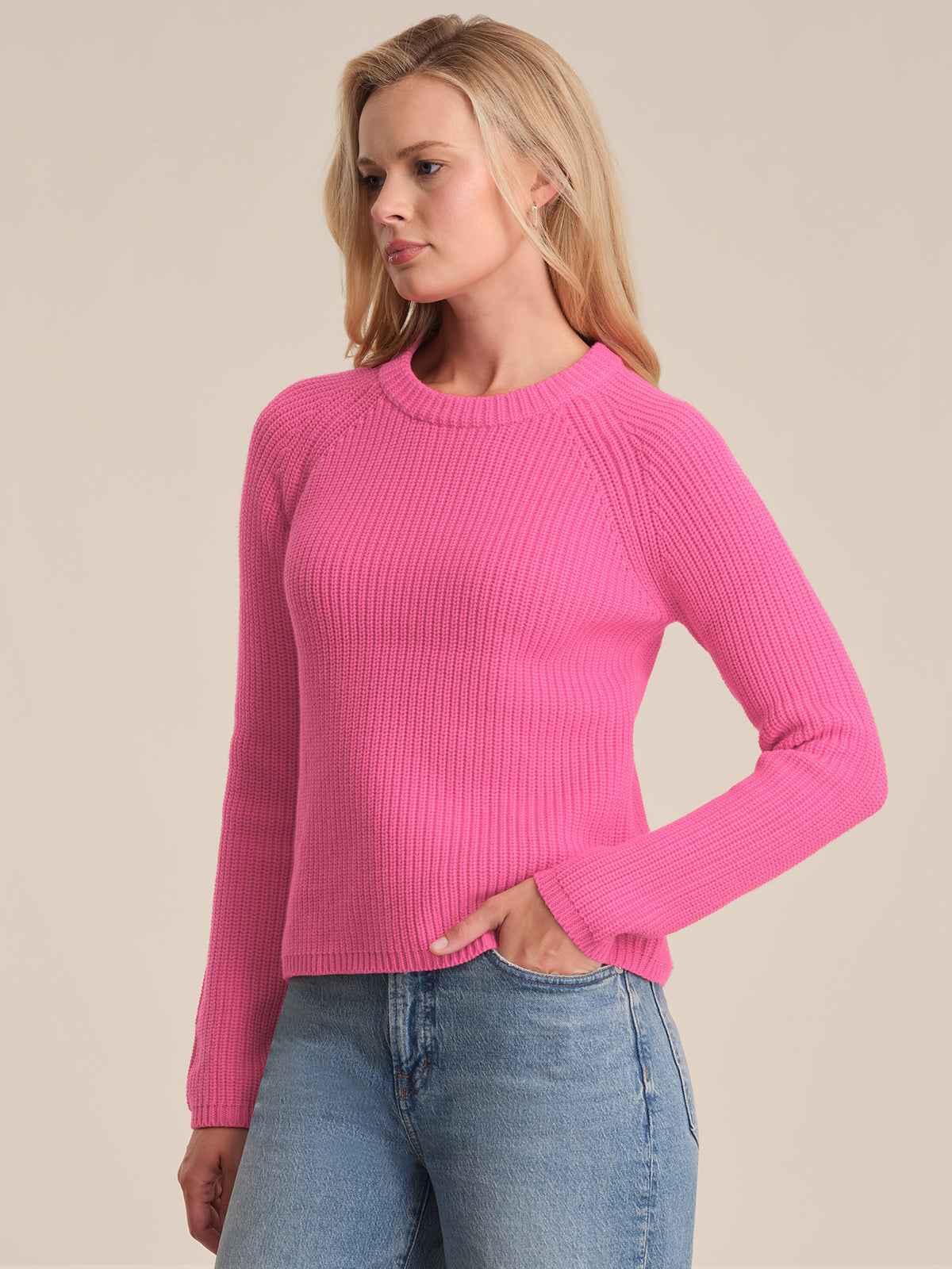 Someone wearing a 525 America bright pink cotton shaker stitch sweater with raglan sleeves and light blue jeans stands against a plain beige background, looking to the side with one hand in their pocket.