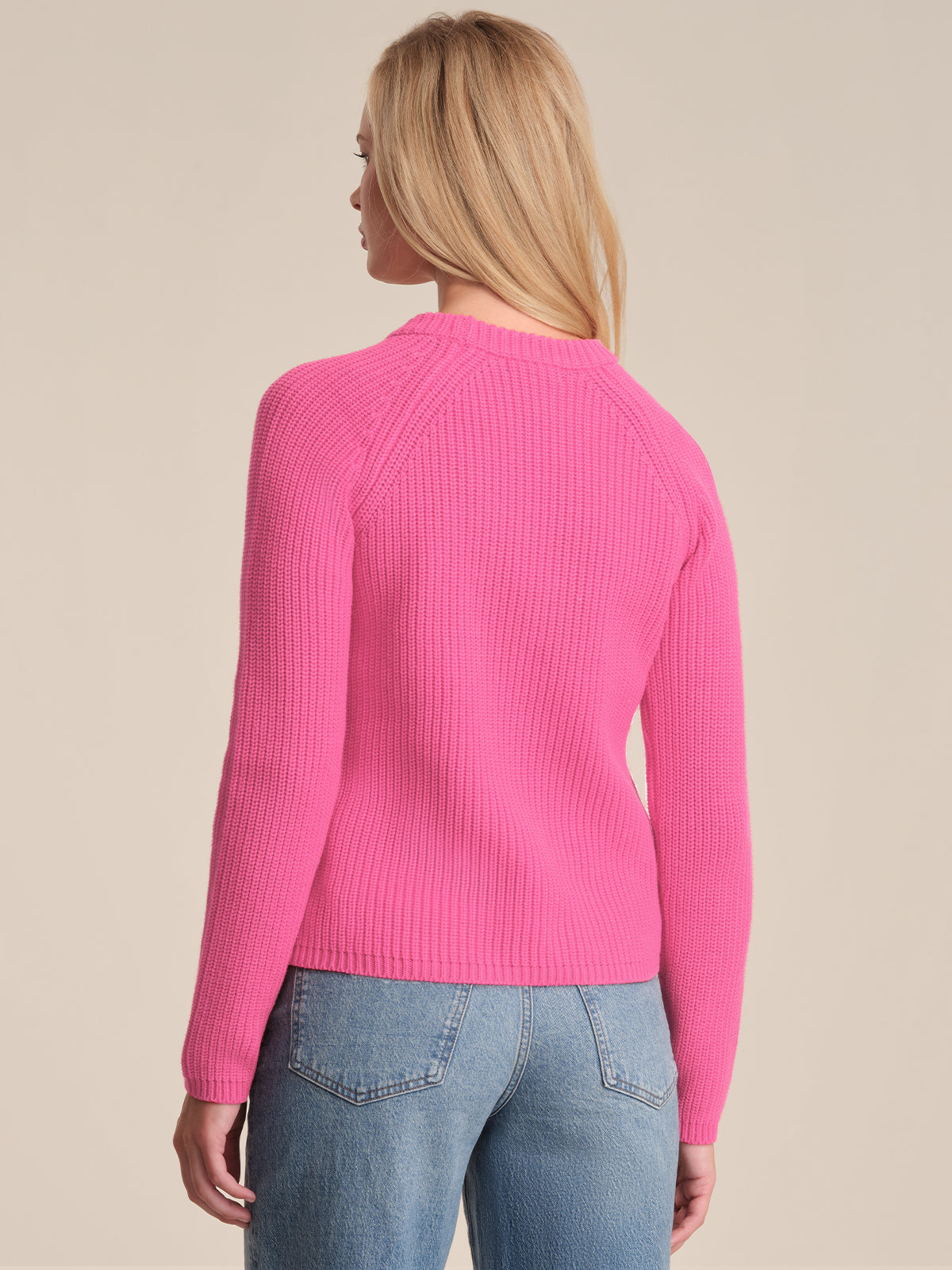 A person with long blonde hair is seen from the back, wearing a bright pink 525 America cotton shaker stitch sweater with a fitted silhouette and blue jeans against a plain, light-colored background.
